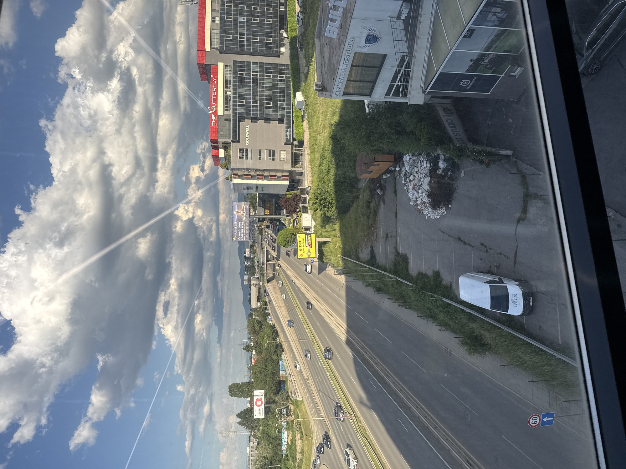 The image shows a view from an elevated position overlooking a busy highway in Sofia, Bulgaria. Notable buildings include "THE BUTTERFLY," "GOLDWELL," and "BULT CAR WRAPPING STUDIO." Vehicles are moving along the highway. A white "IRIS" van is parked along the shoulder. Signs for "PALMS BET" and "efbet" are visible. A speed limit sign indicates 60 km/h. The sky is cloudy. Construction debris is visible on the roadside. The image appears to be taken through a window.