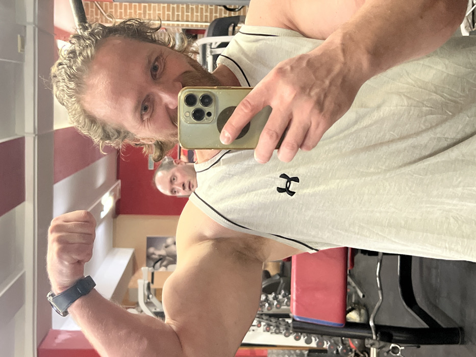 The image displays an interior scene of a gym or fitness center. A man with medium-length, wavy light brown hair, a mustache, and a beard occupies the foreground, looking directly at the camera with a wide smile. He wears a light grey tank top featuring an Under Armour logo on the left chest. His left arm is flexed, prominently displaying a bicep, and a dark smartwatch is visible on his left wrist. He holds a gold-colored smartphone with a multi-lens camera array in his right hand, partially obscuring the lower right side of his face. Reflected in a large mirror behind him, a second man with a receding hairline is visible, looking towards the foreground with a surprised expression, his mouth slightly open. The background, largely reflected, shows gym equipment including weight racks, dumbbells, and a red-padded bench. The walls are partially painted red, and the ceiling contains rectangular light fixtures. The specific location within Saint Petersburg, Russia, is not identifiable from the visual content.