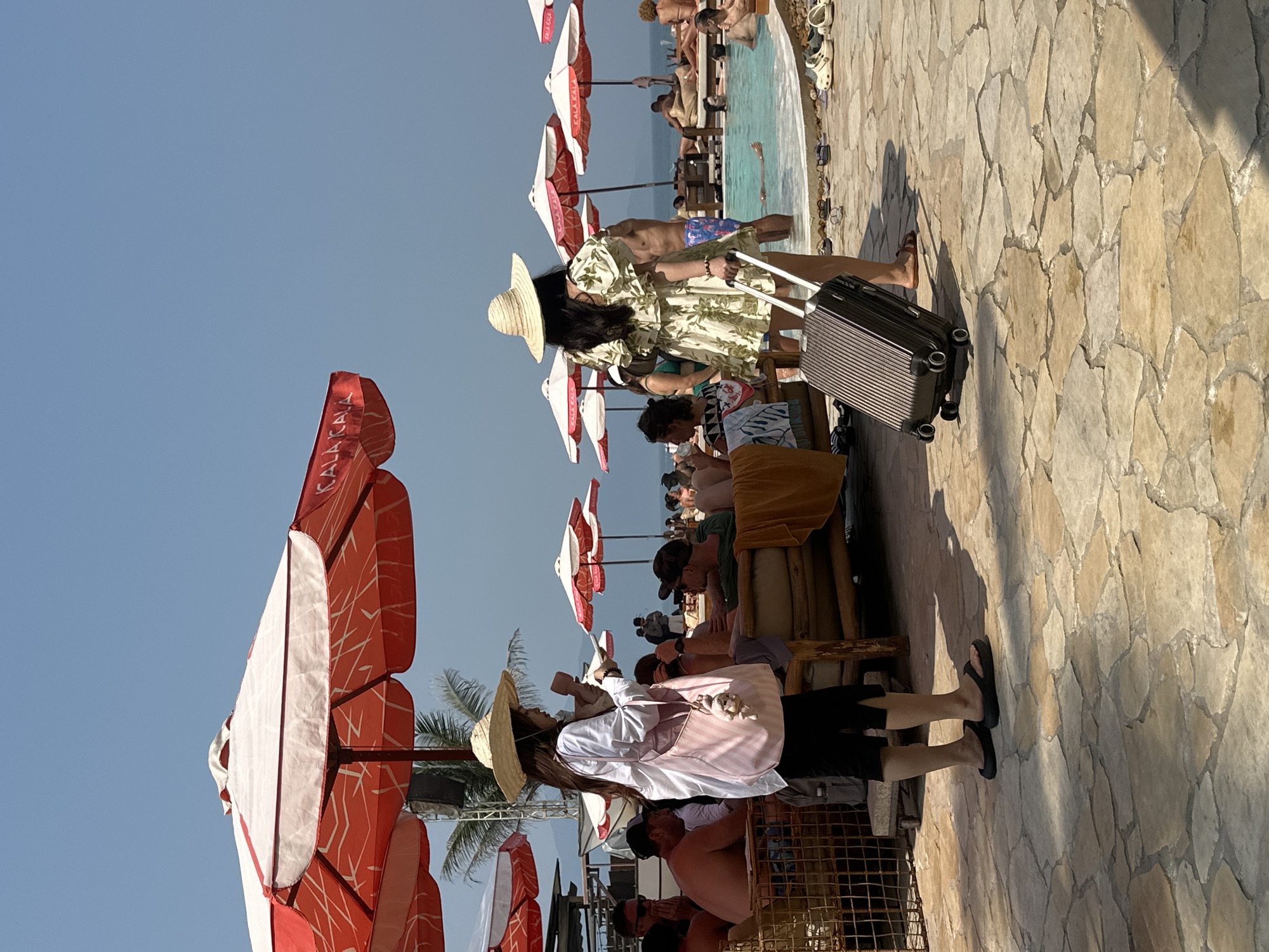 The scene depicts a beach resort in Đà Nẵng, Vietnam. A woman in a floral dress and straw hat is pulling a suitcase along a stone walkway. Behind her, there is a partially visible swimming pool with people in it, bordered by sun umbrellas and loungers occupied by vacationers. In the foreground, another woman in a white shirt, black shorts, and a straw hat is standing among similar beach umbrellas and seating areas. The sky is clear and blue.