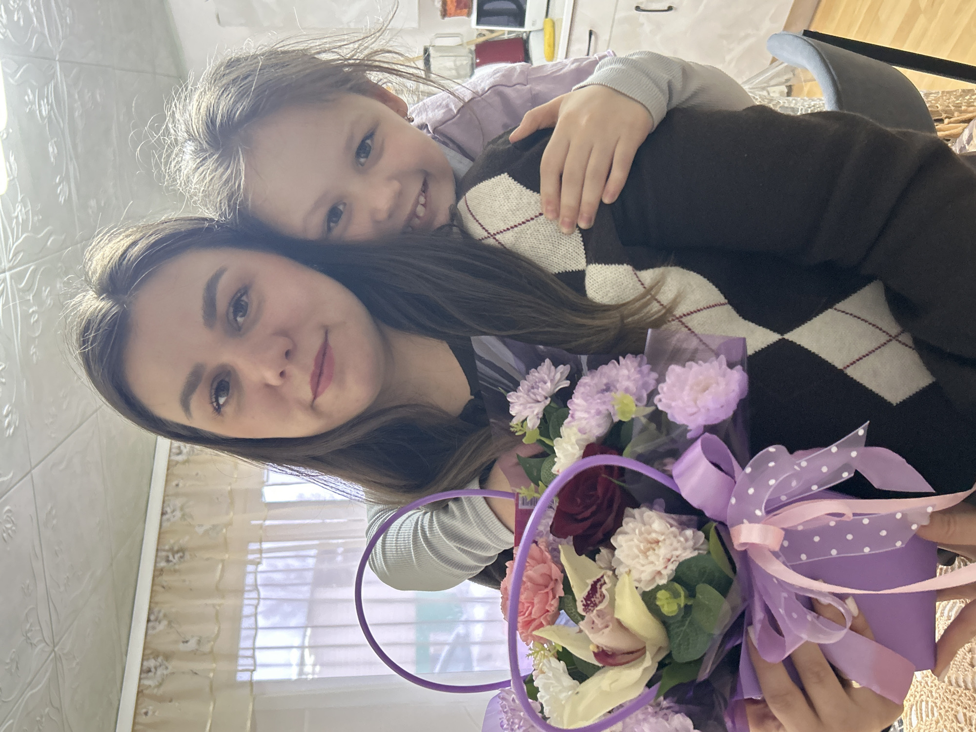 An adult female and a child female are depicted in a portrait-oriented photograph taken indoors in a residential setting within the city of Фоки, Russia. The adult, positioned in the foreground and center-left, has dark brown hair and wears a horizontally striped top under a dark brown argyle sweater with white and red patterns. She holds a purple basket-style floral arrangement containing various flowers, including cream-colored orchids, dark red roses, light pink carnations, and light purple chrysanthemums, adorned with a purple and pink polka-dotted ribbon. Her gaze is directed towards the camera with a slight smile. Behind and to the right of the adult, a child with light brown hair, wearing a light purple long-sleeved shirt, embraces the adult from behind, resting her head on the adult's right shoulder and smiling at the camera.
The background shows a domestic interior, likely a kitchen or dining area. A large window with light-colored sheer curtains is visible on the left, offering a blurred view of an exterior environment. The ceiling features embossed white tiles with a decorative pattern. To the right, white kitchen cabinets, a red blender, a white microwave, and a yellow object (possibly a sponge) on a countertop are discernible. A dark grey chair with a black frame is partially visible on the bottom right. A light-colored, possibly crocheted, tablecloth is visible on a surface in the bottom left foreground.