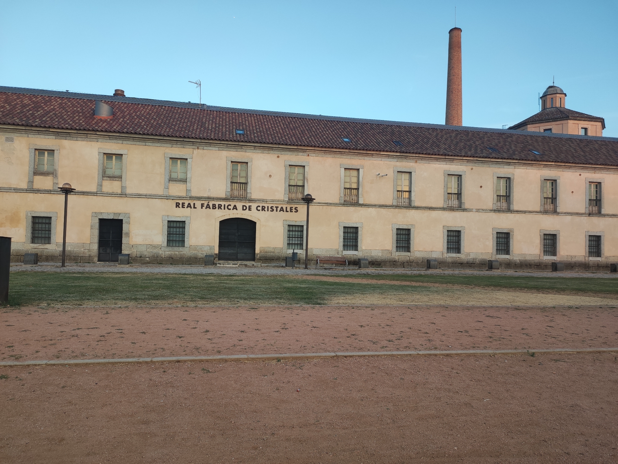 The image shows the exterior of the Royal Factory of Crystal (Real Fábrica de Cristales) in the Real Sitio de San Ildefonso, Spain. The building is a long, two-story structure with a beige facade, a reddish-brown tiled roof, and numerous windows with dark frames. In front of the building is a grassy area, and beyond that, a reddish-brown dirt path. A tall, red brick chimney stands to the right of the building, and a domed structure is visible behind it. The sky is clear and light blue, suggesting it is daytime. There are no people visible in the image.