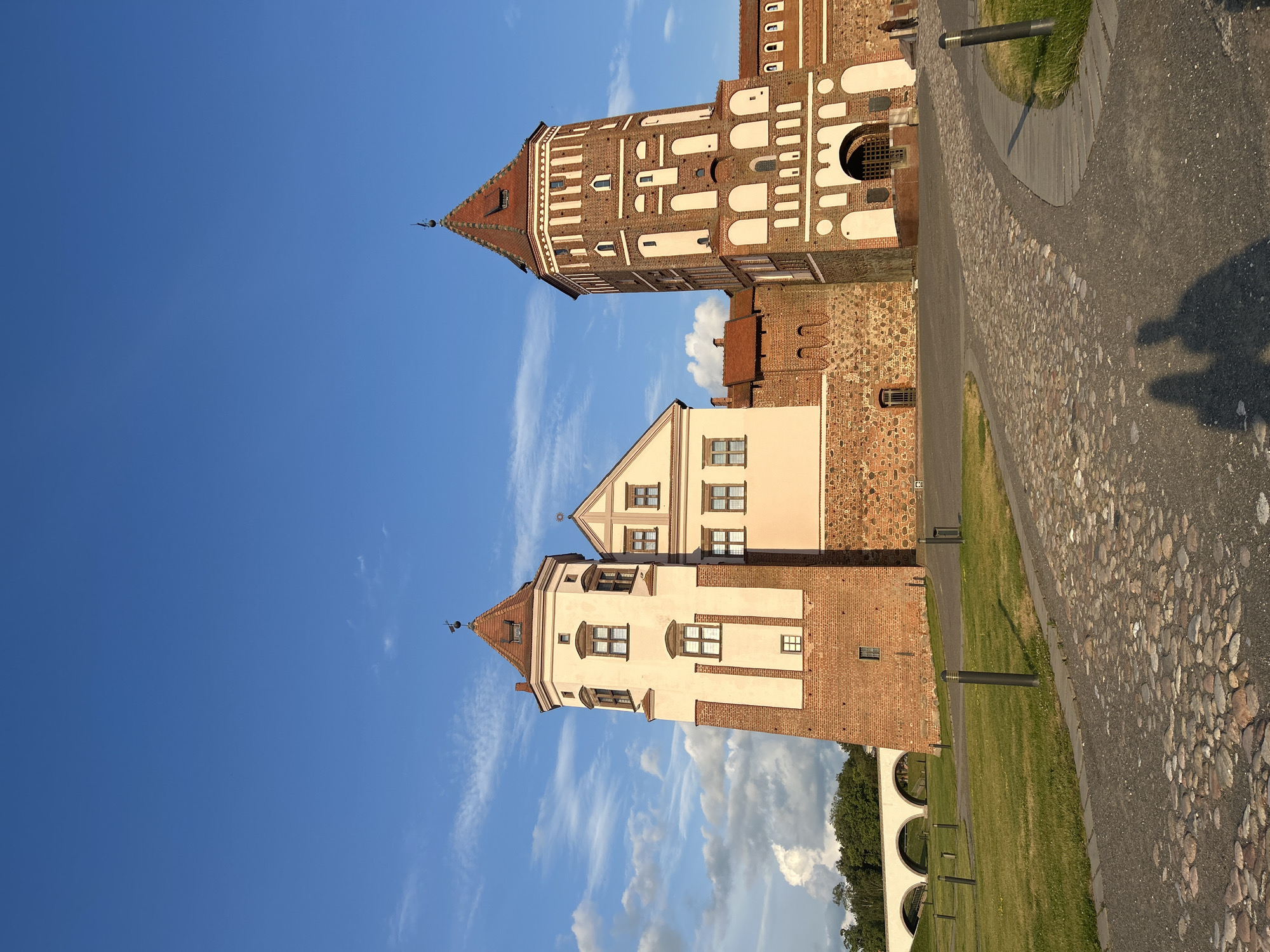The image shows the Mir Castle Complex in the city of Zhukavichy Rural Council, Belarus, on a clear day with a blue sky and scattered clouds. The castle features two prominent towers: one is taller and red brick with white arched detailing and a steep red roof, while the other is shorter, cream-colored, and also has a red roof. Between the towers is a connecting building with a cream facade and red-tiled roofs. The foreground consists of a cobblestone pathway curving through a manicured grassy area with several modern, dark-colored lampposts. In the background to the left, a white arched bridge spans a gap. The overall impression is one of historical architecture bathed in sunlight.