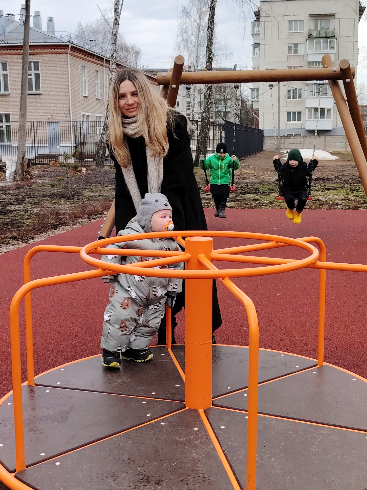 The image shows a playground scene in Chaikovsky, Russia. A woman with long blonde hair and wearing a black coat and light-colored scarf stands near a toddler in a gray animal-patterned snowsuit and gray bear hat who is holding onto an orange merry-go-round. The toddler has a pacifier in their mouth. In the background, two children are on swings. One child wears a green jacket, and the other wears a black jacket and yellow boots. The playground surface is red. Behind the swings is a metal fence and apartment buildings with bare trees in the background. A brick building with light-colored trim is visible to the left of the woman. The sky is overcast.