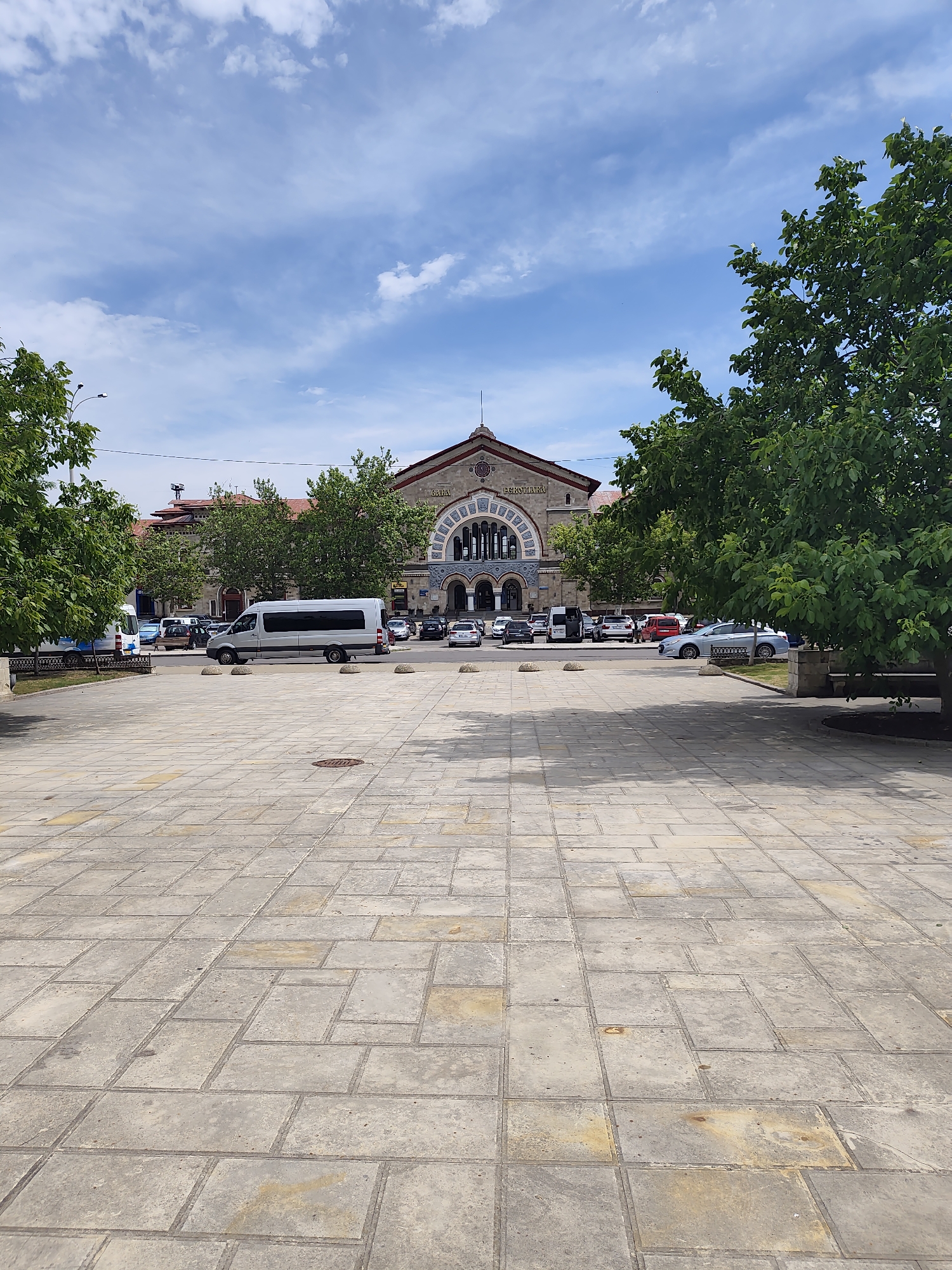 The image shows a view of the Central Market of Chișinău, Moldova, on a sunny day. The foreground is a wide, paved plaza with large square and rectangular stone tiles. Several round, dark manhole covers are visible on the plaza. In the mid-ground, a silver van is parked in front of a row of buildings. Behind the van, there are several cars and a bus. The buildings in the background appear to be the market structure itself, with a large arched entrance and ornate detailing. Green trees are visible on either side of the plaza and behind the market buildings. The sky is blue with scattered white clouds. The scene depicts a public space with vehicles and market architecture, suggesting a bustling urban environment.