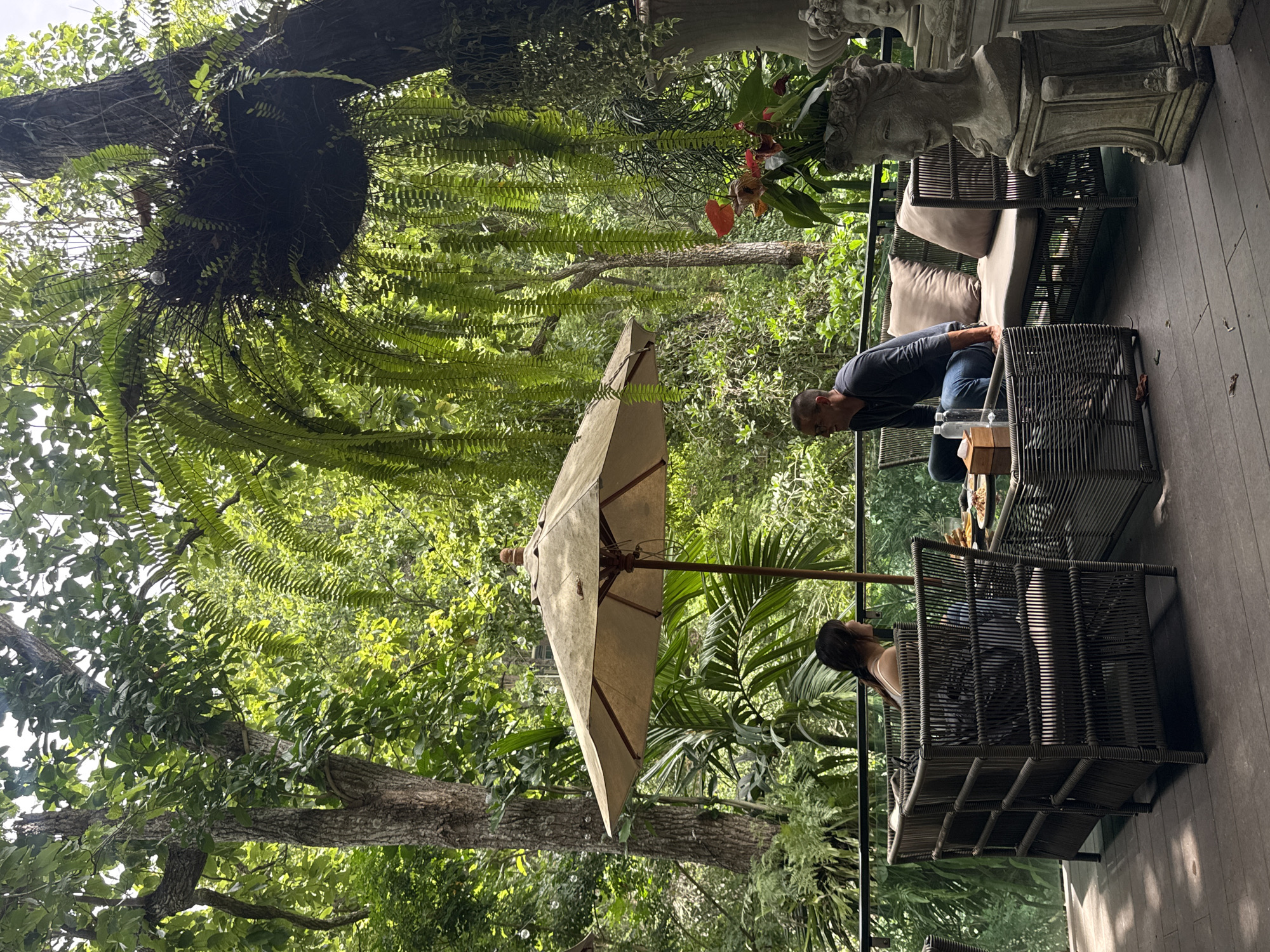 This image depicts an outdoor seating area in Suthep, Thailand, surrounded by lush greenery. Two people are seated at a table under a large umbrella. A man in a dark long-sleeved shirt and blue jeans is seated on a cushioned sofa, looking to his left. A woman with dark hair is seated in a wicker chair, facing away from the camera, also looking to her left. In front of them is a glass-topped table with food and drinks. To the right, a decorative stone bust is visible. The environment is characterized by abundant trees and hanging ferns, suggesting a tropical setting. The seating area is on a wooden deck.