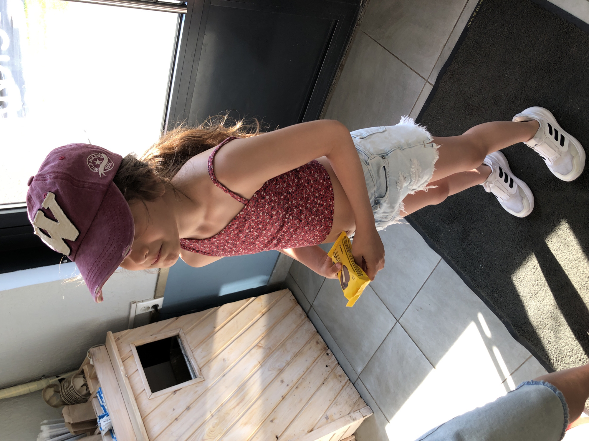A young girl is pictured outdoors, standing. She is wearing a burgundy baseball cap with a white "N" emblem, a floral print tank top, distressed denim shorts, and white Adidas sneakers with black stripes. She is holding a yellow package of what appears to be cookies. The background shows a wooden structure that resembles a small playhouse and a tiled floor. Sunlight casts shadows, suggesting it is daytime. The setting appears to be a residential or outdoor recreational area. There is no discernible text that is legible enough to read.