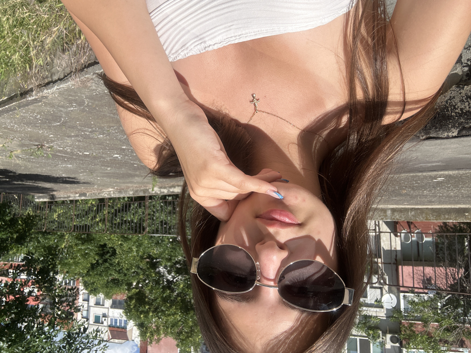 A young woman with long brown hair and sunglasses is taking a selfie outdoors in the city of Shumen, Bulgaria. She is wearing a white strapless top and a gold cross necklace. Her hand is resting on her chin. The background shows a residential building, trees, and a metal fence. The lighting suggests it is a sunny day.