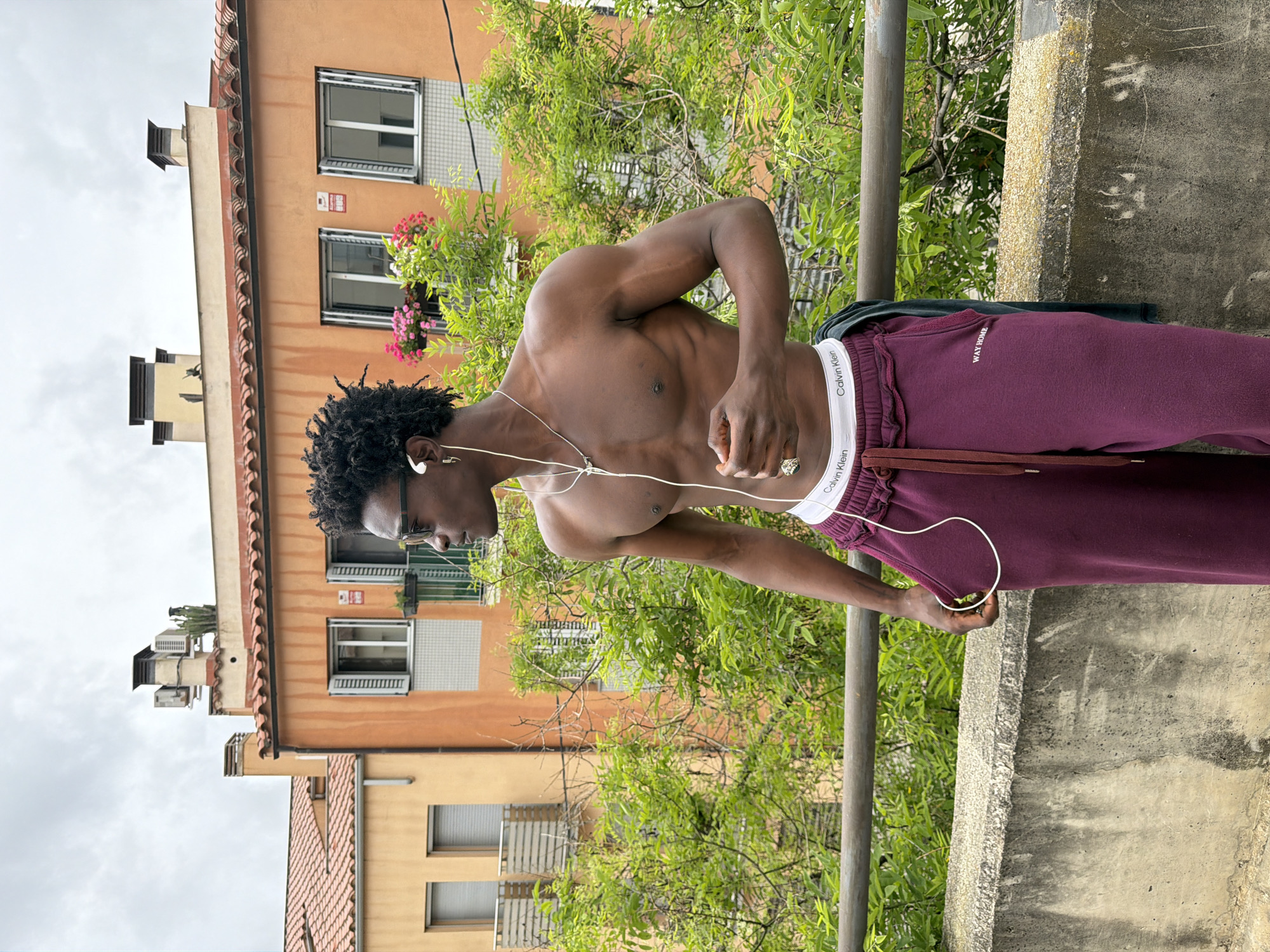 A vertical, full-shot image features a shirtless dark-skinned man with dreadlocks, wearing glasses and maroon sweatpants, listening to headphones while leaning on a railing. The man's sweatpants have a "WAY HOME" logo visible. His white Calvin Klein underwear is exposed above the sweatpants. The backdrop shows an orange building with windows, a tiled roof, and flower boxes. Green foliage is interspersed between the building and the man, while a gray sky looms above. The man is standing on a concrete surface with a textured, weathered appearance. The image appears to be taken outdoors in Sant Andreu De Llavaneres, Spain, based on the architectural style of the building.