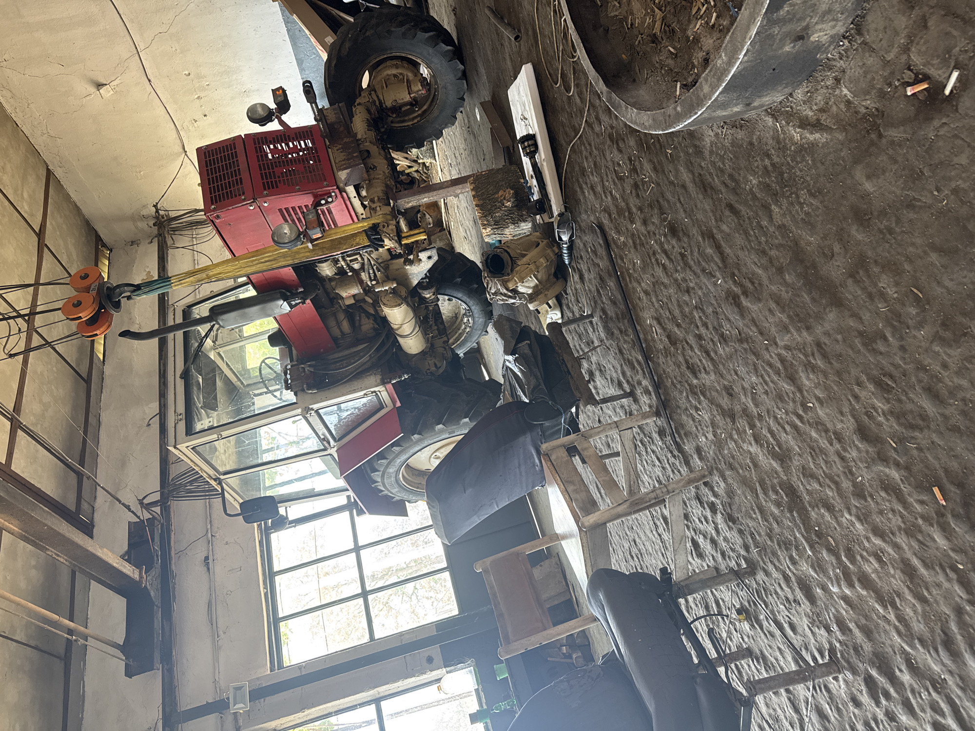 The image depicts the interior of a workshop or garage, likely an agricultural setting, given the presence of a red tractor. The scene is presented from a high angle, looking down onto the dirt floor and various equipment.
The primary subject is a large, red tractor, partially disassembled, with its cab visible on the left side of the frame. It features robust tires, an exposed engine or transmission area, and a large exhaust pipe. Attached to the front of the tractor is a yellow strap connected to a hydraulic crane mechanism with orange pulleys, suggesting it's either being used for lifting or is part of the tractor's auxiliary equipment. Red grates or panels, possibly housing a generator or other machinery, are visible on the tractor's structure.
The floor is a rough, packed earth surface, scattered with various debris including numerous cigarette butts, particularly noticeable around a large, dark metal ring or tire in the foreground right. Nearby, a wooden plank, a log, and what appears to be a disassembled mechanical component (possibly a gearbox or transmission) along with a drive shaft, are laid out, further indicating maintenance work.
The workshop walls are light-colored, showing signs of wear and cracks, with exposed metal beams, conduits, and electrical wiring. Natural light enters through large windows with multiple panes, illuminating the space and suggesting it is daytime. A small white light fixture is mounted on a wall. Wooden chairs or stools are also visible, some appearing rustic. The overall impression is one of a functional, lived-in workspace for heavy machinery repair and maintenance in a rural Bulgarian context.