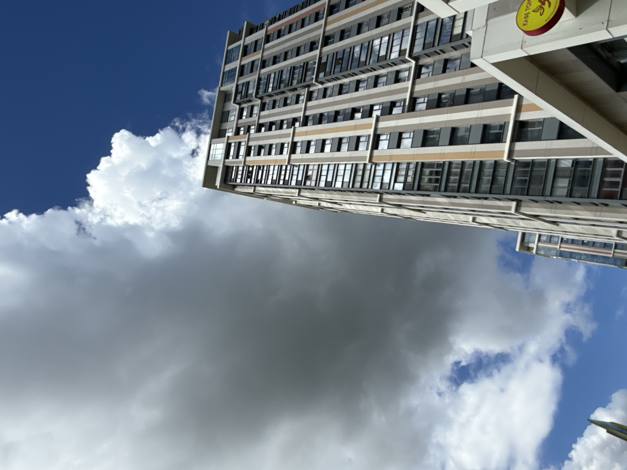 The image depicts a multi-story building in Moscow, Russia, seen from a low angle against a sky filled with white and dark clouds. The building has a repetitive pattern of windows and beige-colored horizontal bands. A sign with Cyrillic text, partly obscured, is visible on the building's edge. No people are visible in the image.