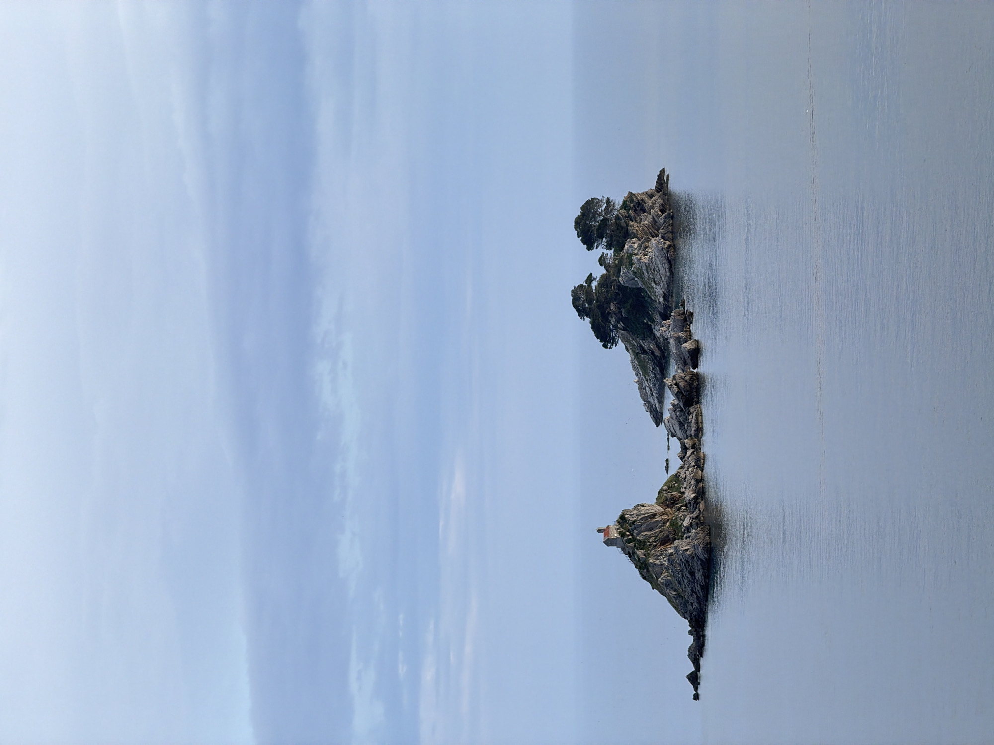 The image shows two small rocky islands in the sea. The island closer to the viewer has a small structure on it, while the other island is topped with trees. The sky is overcast and the water is calm. The location is Petrovac na Moru, Montenegro.