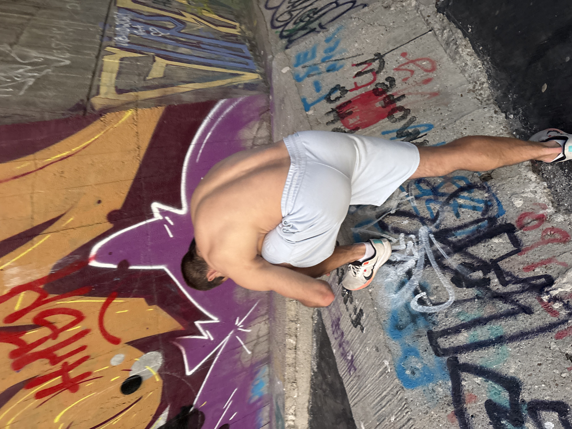 A shirtless male is squatting on a concrete embankment covered in graffiti. The man is wearing light blue athletic shorts and white sneakers with orange and black accents. The embankment is part of an urban environment, possibly under a bridge or overpass, with a graffiti-covered wall behind him. The graffiti on the wall features bold, colorful lettering and designs in red, yellow, purple, and blue. The ground is also covered in various tags and markings. The lighting suggests it is daytime, and the overall scene appears to be an outdoor, public space.