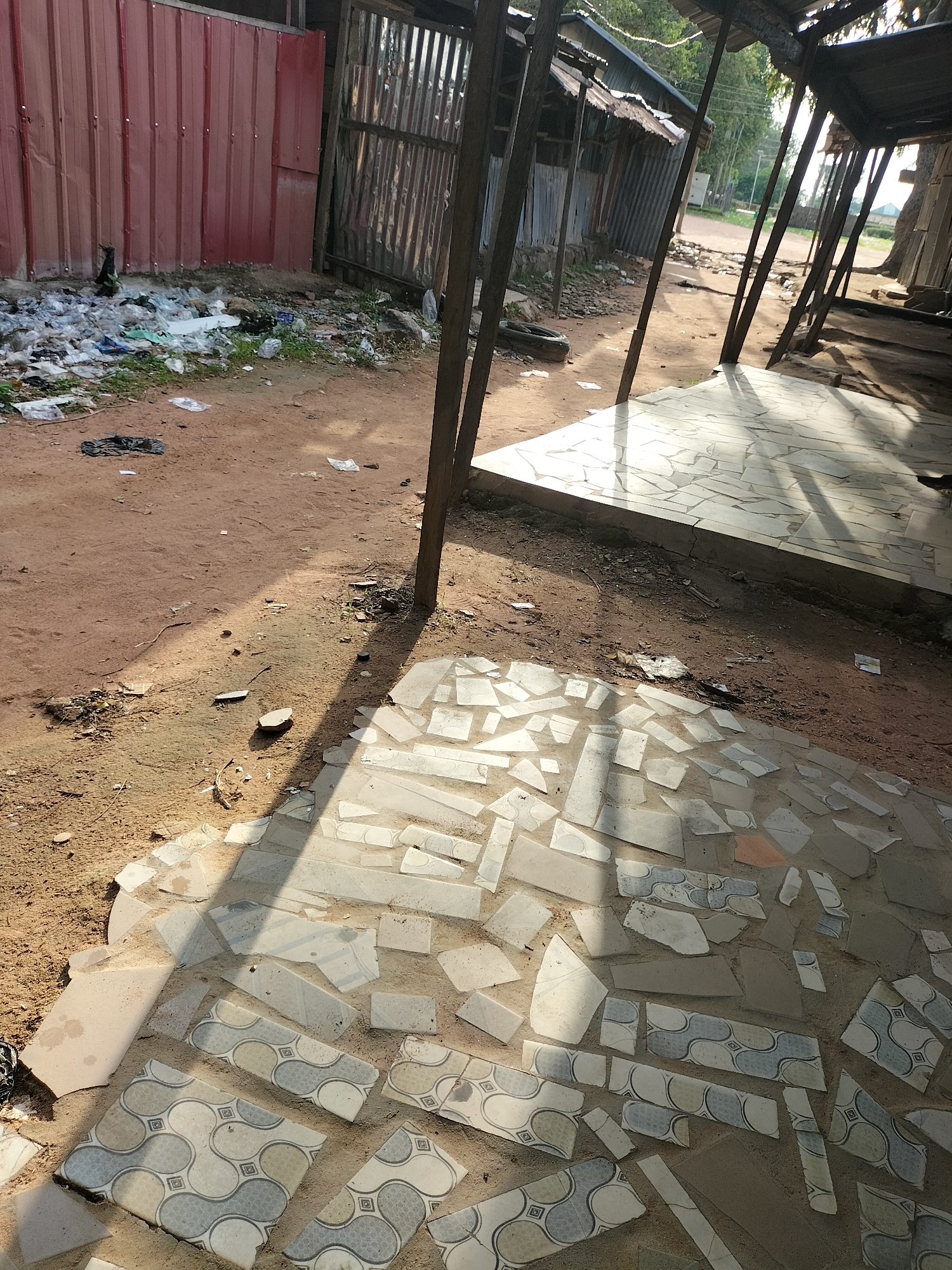 The image depicts an outdoor scene in Jalingo, Nigeria. In the foreground, a patch of ground is covered with broken tiles, some with a patterned design featuring blue and grey wavy lines within squares. This tiled area is adjacent to a dirt path with scattered debris, including plastic waste and what appears to be a black plastic bag. A thin shadow stretches across the tiled area, suggesting it is daytime.
In the mid-ground, a corrugated metal fence, painted red on the left and silver on the right, separates the foreground from a more cluttered area. This area features more debris, including plastic bottles, bags, and other unidentifiable refuse, mixed with some sparse vegetation. Beyond the fence, a row of small structures with corrugated metal roofs and wooden supports is visible. The structures appear weathered and are aligned along a dirt road.
The background shows trees and vegetation, indicating a semi-urban or rural setting. The overall impression is one of a somewhat neglected or impoverished area, possibly a market or residential periphery. No people are visible in the image, and no specific actions or interactions are depicted. The lighting suggests it is either early morning or late afternoon, with the sun casting long shadows. The scene is captured from a slightly elevated, outward-looking perspective.
