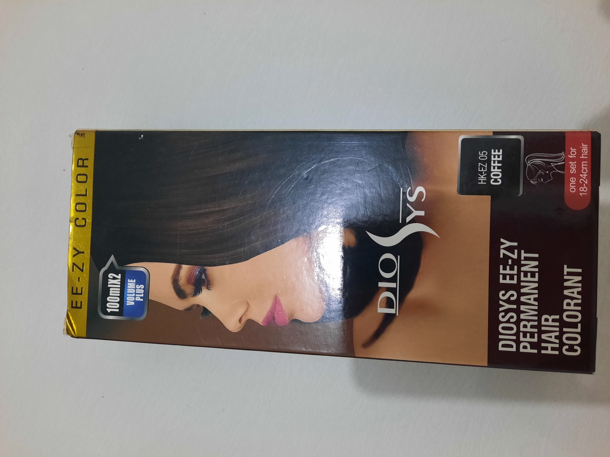 The image shows the front of a boxed hair dye product. The box is predominantly dark brown and features a stylized graphic of a woman's face in profile, with dark hair and makeup. The branding includes "EE-ZY COLOR" at the top in yellow text, and "DIOSYS" in large white lettering. There are also callouts for "100mlX2 VOLUME PLUS," a color designation "HK-EZ 05 COFFEE," and a note indicating "one set for 18-24cm hair." The product appears to be positioned on a light-colored, possibly white, surface. The lighting suggests an indoor environment, with some slight glare on the box's surface. There is no indication of the city of Lhokseumawe, Indonesia, in the image or on the product packaging.