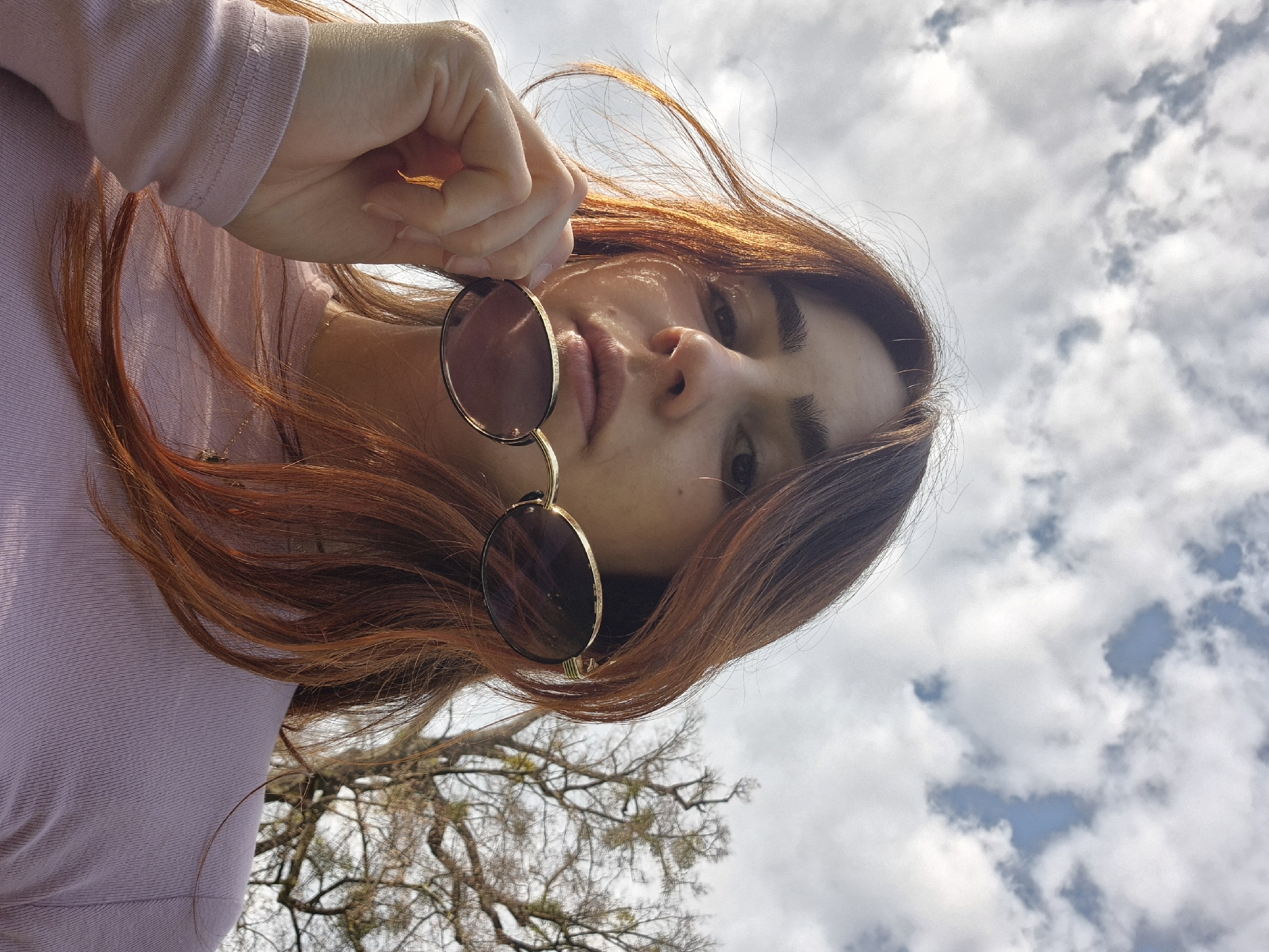 A low-angle photograph captures a female subject outdoors in Strasbourg, France. The subject, with long, reddish-brown hair, wears a light pink, long-sleeved ribbed top and a delicate gold chain necklace. Her right hand holds a pair of round, gold-framed sunglasses with dark lenses below her chin, positioned slightly to her left. The brand "BENJAMIN" is legible on the left temple of the sunglasses. She looks directly at the camera with a neutral expression. Sunlight illuminates the left side of her face. The background consists of a partially overcast sky with white and grey clouds, revealing intermittent patches of blue. To the right, bare tree branches, some with green foliage, are silhouetted against the sky.