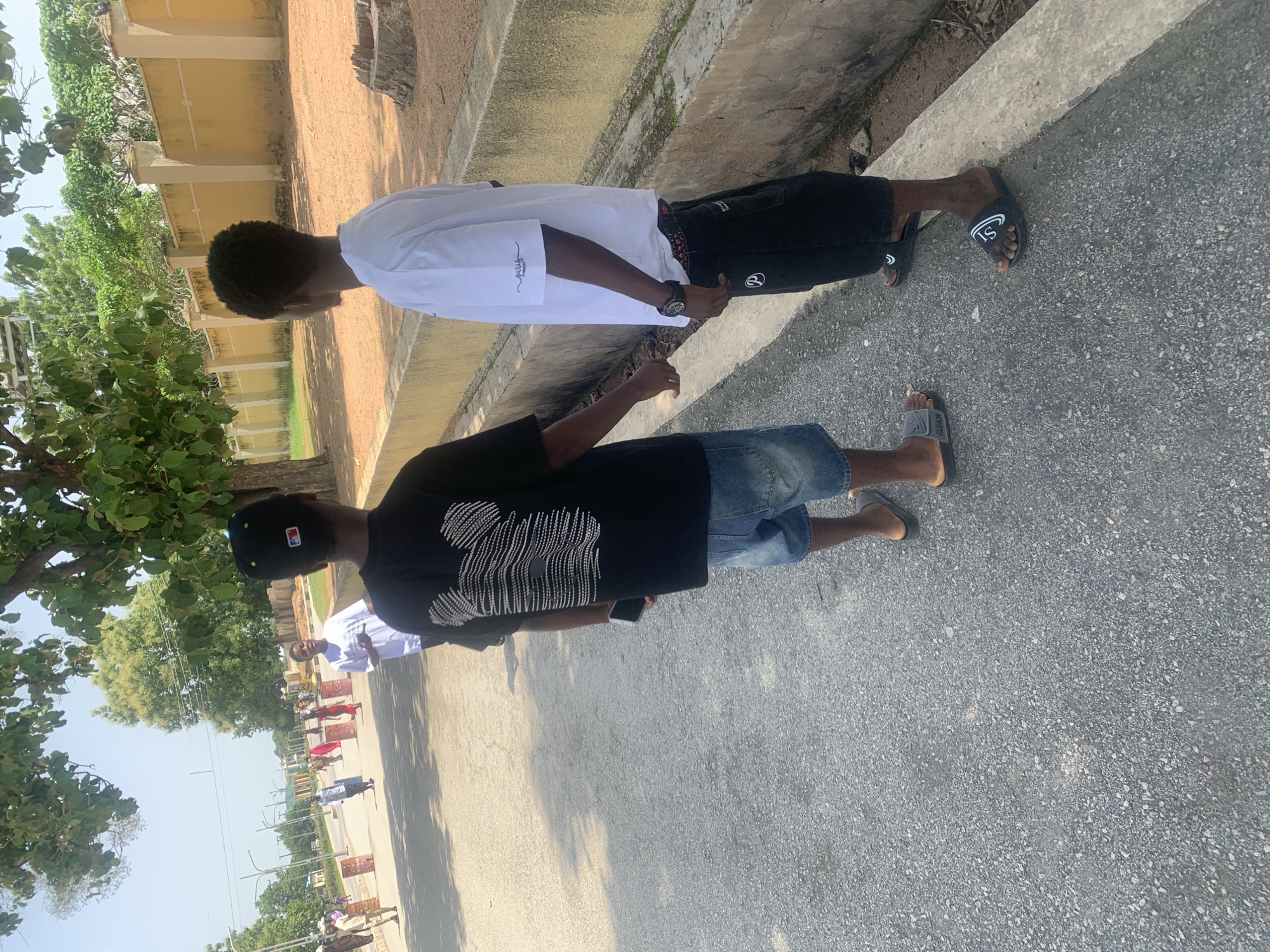 Two young men are standing on a paved road in what appears to be Jalingo, Nigeria. The man on the left is wearing a white short-sleeved shirt with subtle embroidery on the sleeve, black shorts, and black slides with a white logo. He has a black watch on his left wrist. The man on the right is wearing a black t-shirt with a white graphic design on the back, denim shorts, and grey slides with a white stripe. He is also wearing a black baseball cap.
The scene is outdoors, with a building or wall on the left side of the image and a tree with green foliage providing shade. The weather appears to be sunny, casting shadows on the ground. The time of day is likely daytime, given the bright lighting. In the background, a street can be seen with several other people and some roadside barriers, suggesting an urban or semi-urban environment. The overall mood seems casual and relaxed. There is no discernible text that is legible enough to read.