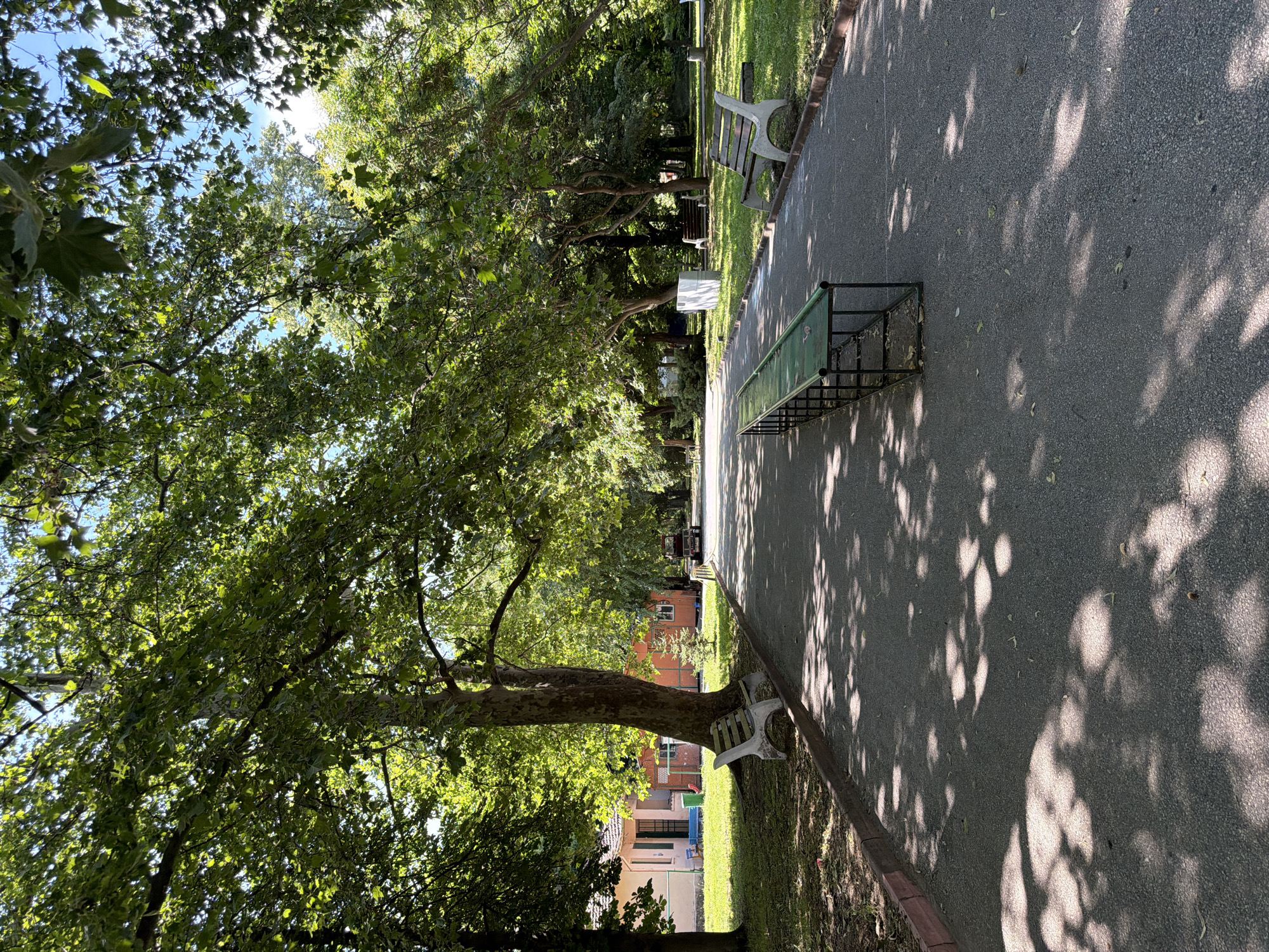 The image depicts a paved pathway in an outdoor setting, likely a park or recreational area. Large trees with dense green foliage line both sides of the path, casting dappled shadows on the ground. Sunlight filters through the leaves, creating a bright and airy atmosphere.
On the left side of the path, a white bench with a slatted wooden seat is situated on a grassy area. Further down the path on the left, a light-colored building with a pinkish facade and a red-tiled roof is visible, accompanied by smaller structures.
In the center of the path, a long, green, metal bench with a dark frame is placed. On the right side of the path, another white bench with a slatted wooden seat is visible, similar to the one on the left. Beyond the benches, the area consists of well-maintained grassy lawns and more trees. In the distance, a dark-colored vehicle is parked on the path.
The scene suggests a peaceful and well-kept public space, suitable for relaxation and recreation. The overall impression is one of a serene, sunlit day in a natural environment within an urban or suburban context. This scene is consistent with the appearance of the park grounds at the Georgi Benkovski Sports Complex in Pazardzhik, Bulgaria, which features a similar layout with tree-lined pathways, benches, and recreational facilities.