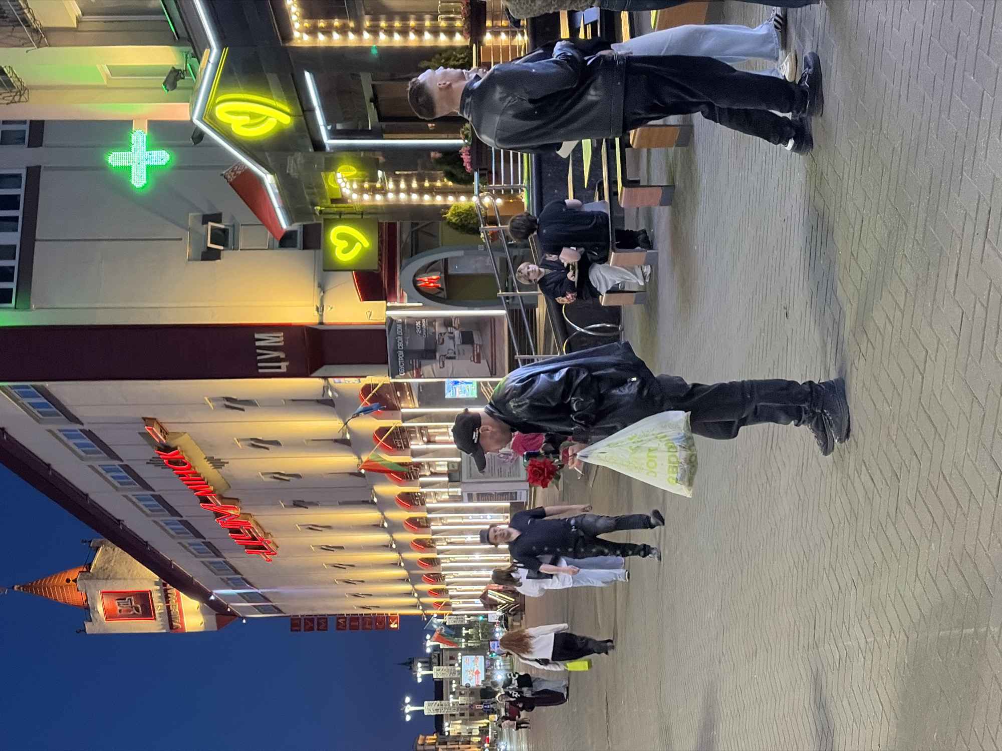The image shows a street scene in Minsk, Belarus, at dusk. The building on the left prominently features the red neon sign "МАГДЕБУРГ ЦЕНТР" along with additional lighting. Further down the street, a building with a neon green cross indicating a pharmacy and another with "ЦУМ" signage is visible. A row of people are walking on the sidewalk, some carrying bags and one holding red flowers. Some are sitting on benches. The pavement is made of interlocking blocks. The sky is transitioning from daylight to night. Artificial lights from shops and street lamps illuminate the area.