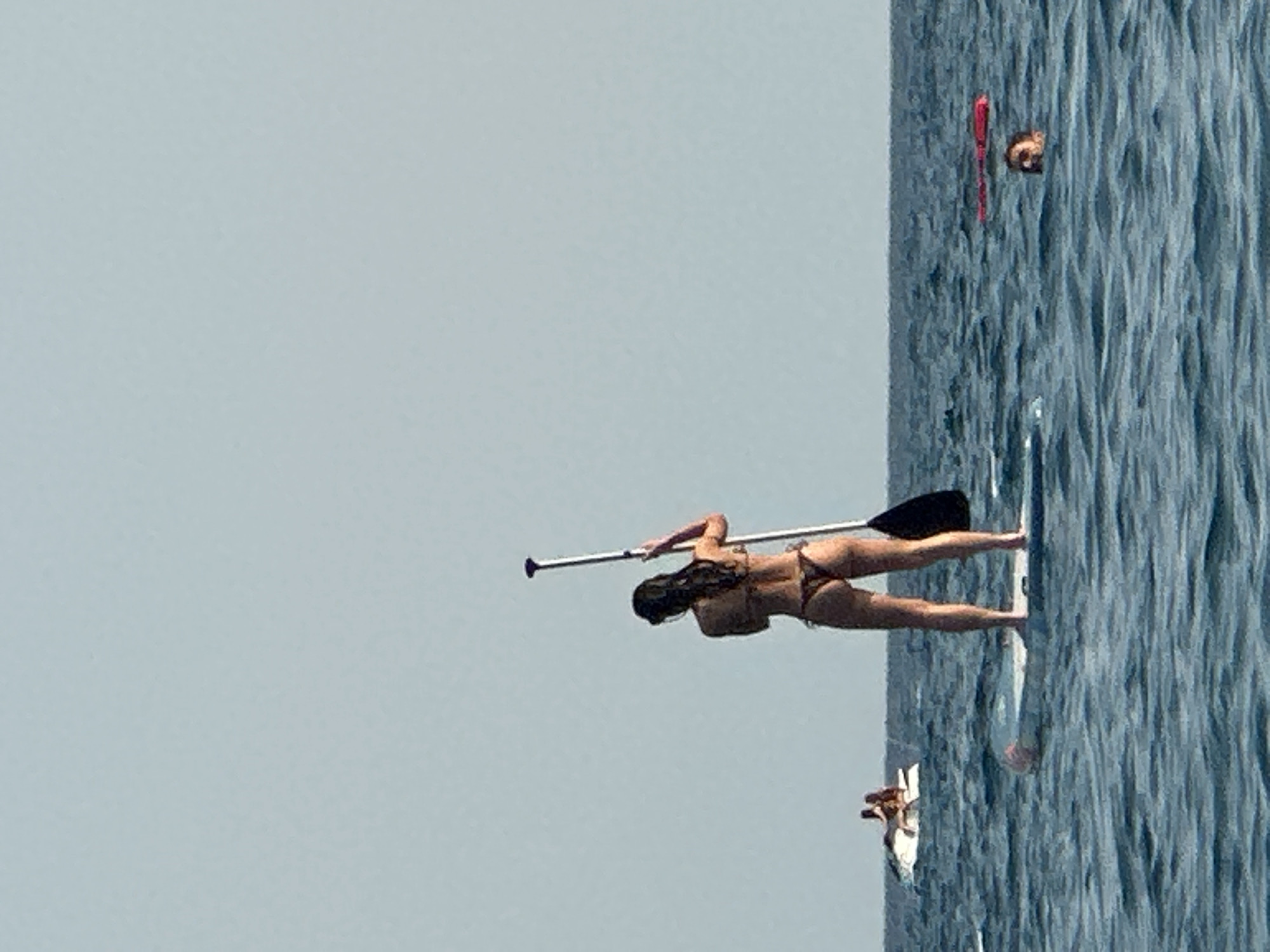 In the city of Boreti, Montenegro, a woman stands on a paddleboard in the calm, blue sea, holding a paddle. She is wearing a dark bikini. In the background, two people are visible on a jet ski, and another person's head is visible above the water with a red float nearby. The sky is clear and pale blue.