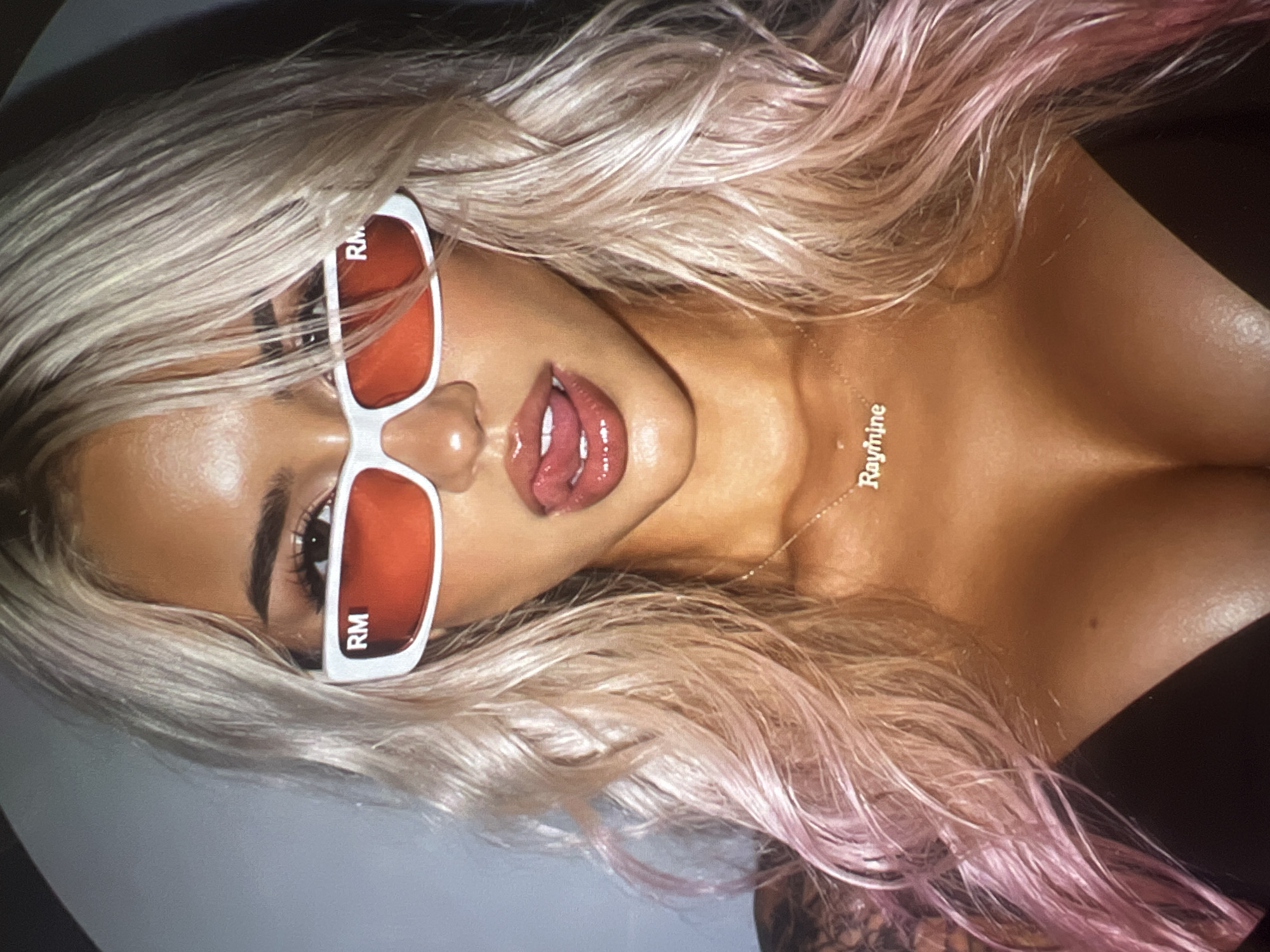 A close-up, head-and-shoulders studio portrait captures a woman with blonde hair, dyed with pink highlights, and red-tinted rectangular sunglasses. She is looking directly at the camera with her tongue playfully sticking out. A silver necklace spelling "Raymine" rests on her collarbone. The background is a plain, light gray. The image does not contain any identifiable landmarks or elements that would suggest a location in Minsk, Belarus.