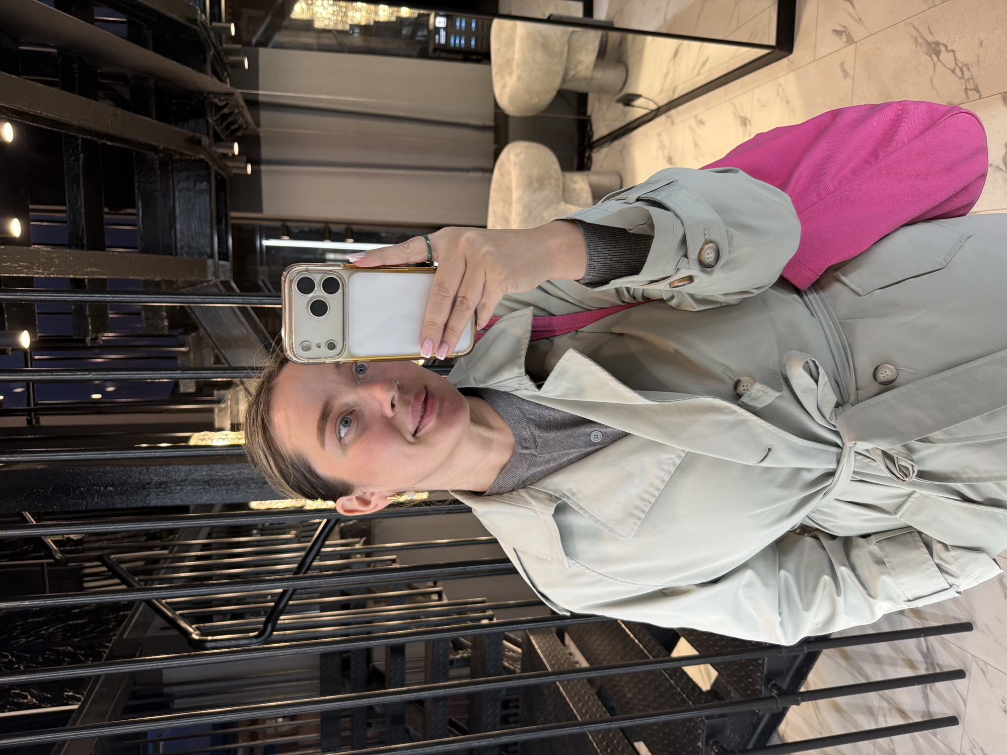 A woman is taking a selfie in what appears to be an urban setting, potentially a building with unique architectural features. She is wearing a light-colored trench coat over a gray top and seems to be sitting on a pink object or cushion. The background includes what looks like a decorative staircase or metal structure and a reflection in a mirror showing a modern interior with marble flooring and light fixtures. Considering the architecture and interior design elements visible, and the assumption that this is Moscow as per the instructions, a plausible location could be a high-end shopping mall or a modern office building.