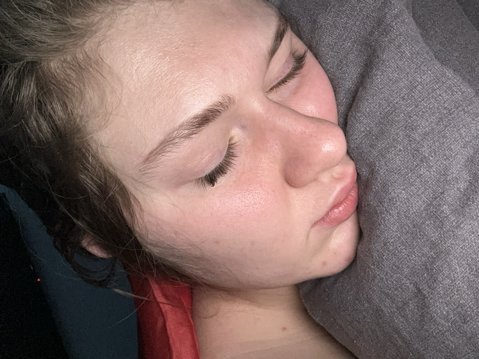 A close-up shot presents a person sleeping peacefully, their face pressed against a gray pillow. The individual has fair skin, visible pores, and well-defined eyebrows. Closed eyes showcase long, dark eyelashes. Their lips are slightly parted. The scene is bathed in natural, muted light, casting gentle shadows. The image conveys a sense of rest and tranquility.