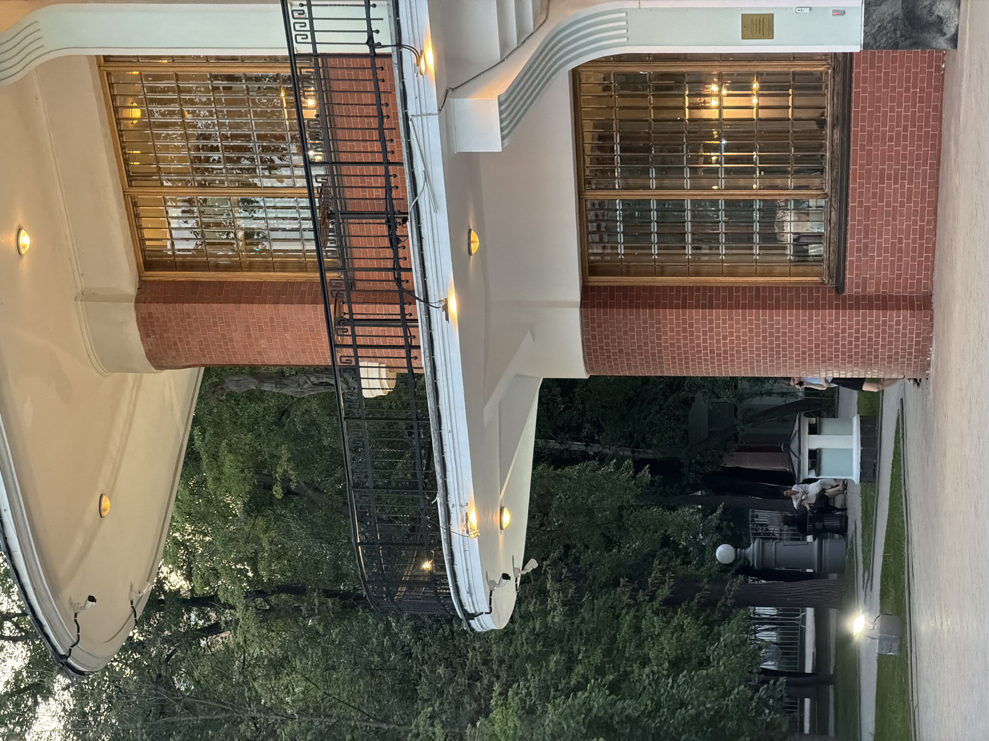 The image captures a building with distinctive architectural features, including a curved balcony with a black metal railing, large windows with a grid pattern, and red brick accents. The structure is illuminated by small lights along the balcony. In the background, a park-like setting is visible with lush greenery, suggesting the location might be near a recreational area. The presence of streetlights, benches, and people engaged in outdoor activities like sitting and socializing implies it's a public space. The overall setting could be near the Melnikov House in Moscow, Russia, given the architectural similarities.