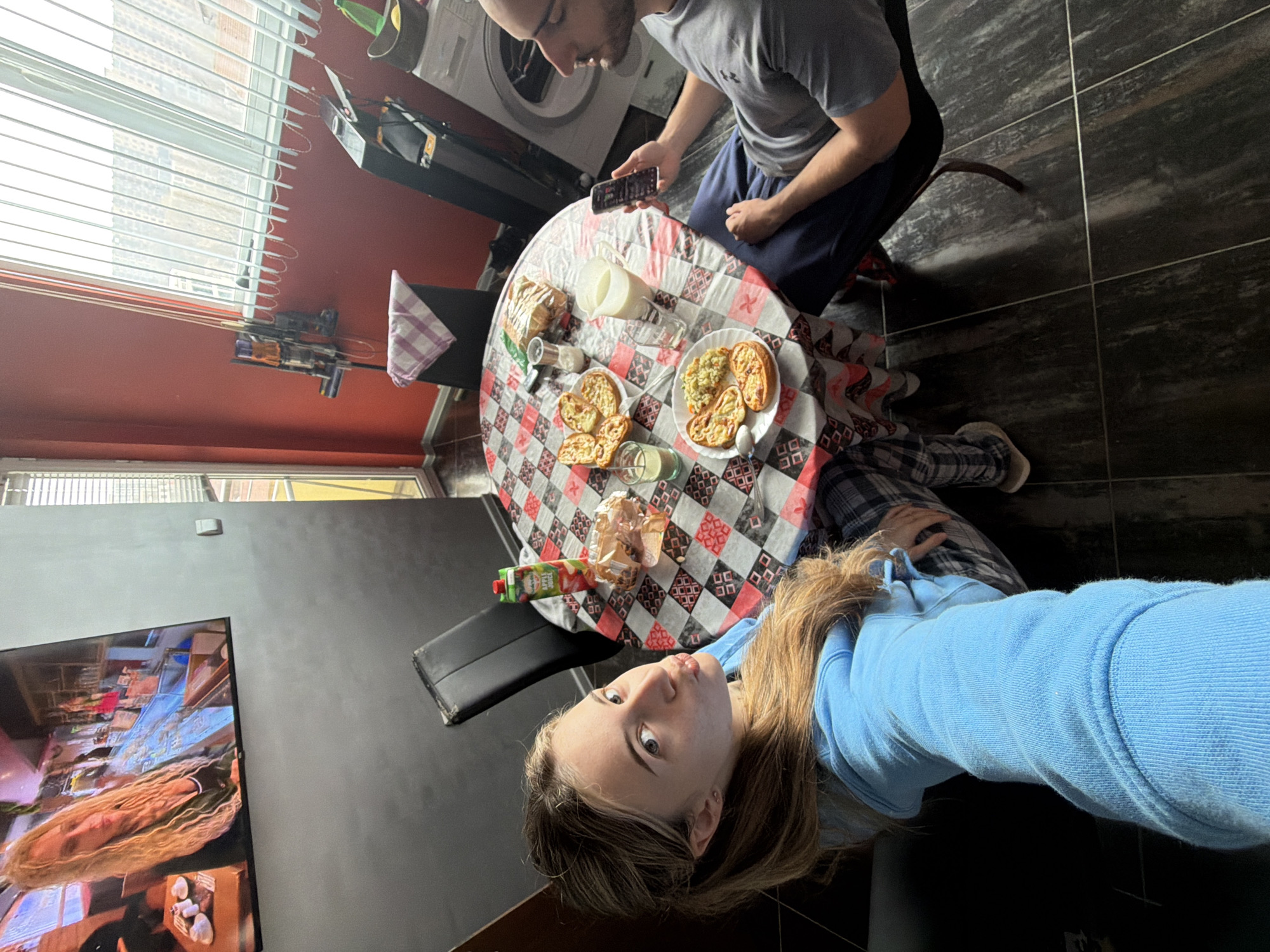 The image depicts an indoor domestic scene, likely a kitchen or dining area, located in Plovdiv, Bulgaria.
In the foreground, a female individual with long, light brown hair, wearing a light blue hooded sweatshirt and plaid pajama pants, captures a selfie, extending her left arm. Her lips are pursed, and she gazes directly at the camera. She is seated at a round table covered with a red, white, and black checkered tablecloth.
To the right, a male individual is seated at the same table, viewed in profile, wearing a gray t-shirt and dark blue pants. He holds a smartphone in his right hand and appears to be viewing its screen.
The table is laden with food and beverages. Items include two plates of savory pastries (some with a green topping), one plate also containing a portion of rice or couscous, a clear pitcher filled with milk, two drinking glasses (one containing milk, the other a green liquid), a small carton of "Bravo" apple juice, and a clear plastic bag containing multiple bread rolls. Salt and pepper shakers, along with a spoon, are also on the table.
The room features dark gray tiled flooring. The wall to the left is painted gray, where a flat-screen television is mounted, displaying a woman with long blonde dreadlocks or braids. The wall to the right is painted red. A Dyson stick vacuum cleaner is mounted on the red wall. A white washing machine and a black treadmill or similar exercise equipment are positioned on the floor against the red wall. A large window fitted with white vertical blinds offers an exterior view of rooftops and buildings. Adjacent to the window is a glass door or another large window, also with vertical blinds, revealing yellow exterior elements. A black dining chair with a small, checkered kitchen towel draped over its back is visible near the table.