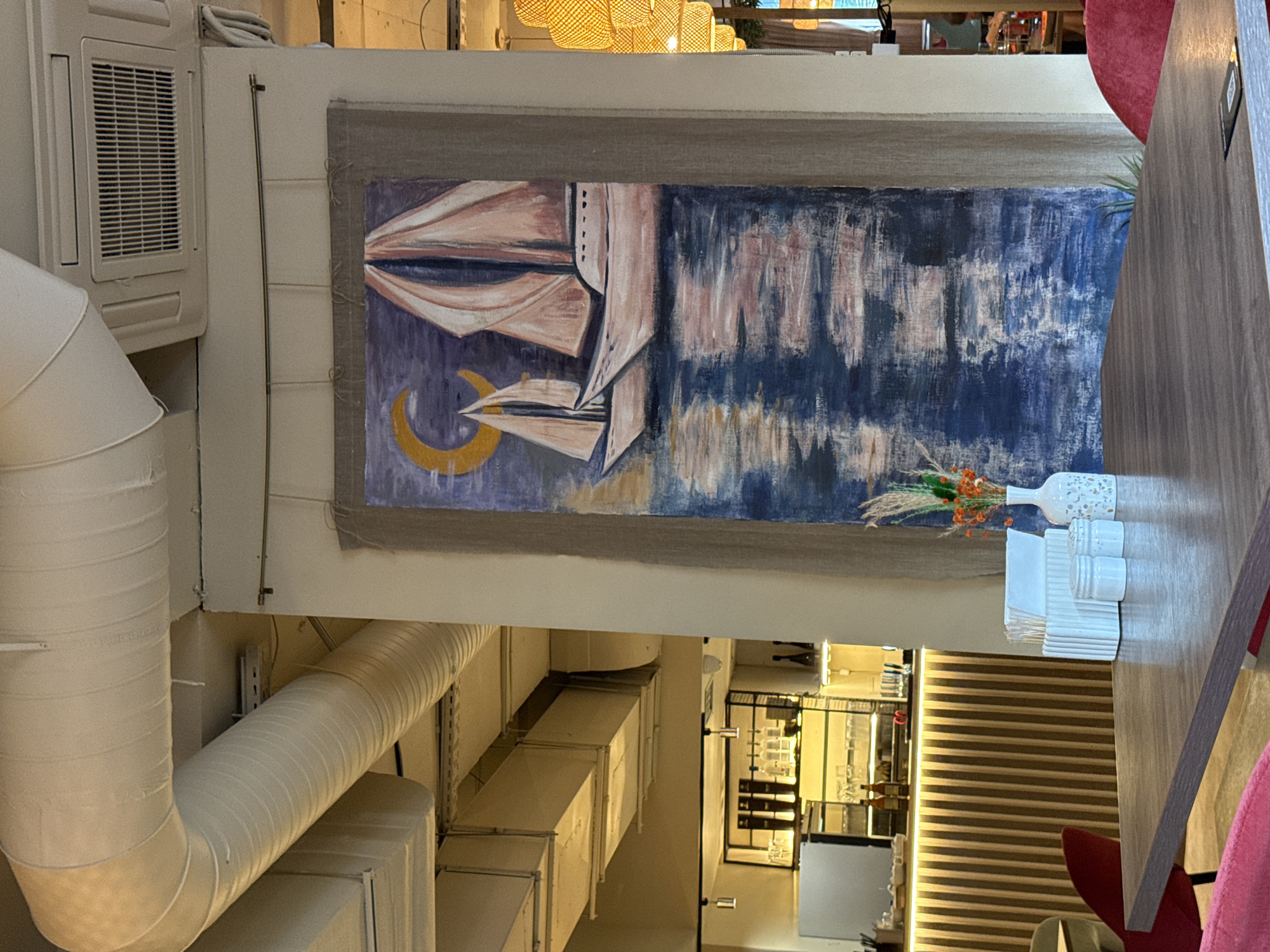 This is a horizontal, eye-level, indoor shot of a restaurant interior. The scene is dominated by a large, abstract painting hanging on a light-colored wall. The painting features a dark blue and white textured background with abstract shapes in pink and beige. In the lower left of the painting, a golden crescent moon is visible. To the right of the painting, a wooden table is set with a white vase holding dried flowers, two small ceramic salt and pepper shakers, and a stack of white napkins. Part of a wooden countertop and a red chair are also visible on the right side of the frame. In the background, above the painting, a series of woven light fixtures illuminate the space. Visible to the left of the painting is a white air conditioning unit with a horizontal vent. Large, cream-colored ventilation pipes extend from the ceiling downwards, dominating the left side of the image. Further in the background, through an opening, a bar area is visible with shelves, bottles, and a refrigerated display. The lighting suggests it is either daytime or evening. The overall impression is that of a modern, art-focused dining establishment. There is no visible text in the image.