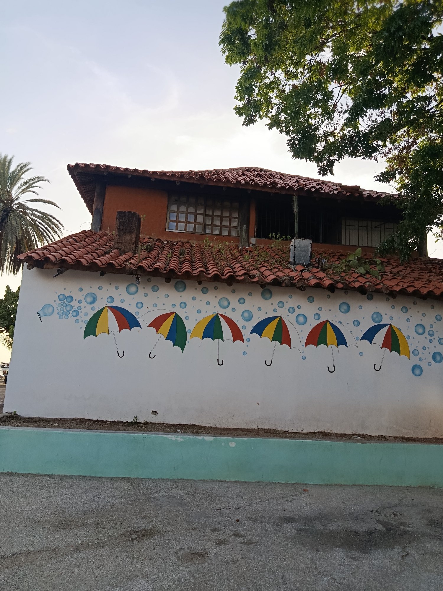 A side view of a white building in Juan Griego, Venezuela, features a mural of colorful umbrellas and blue bubbles painted on its exterior wall. The building has a red tiled roof and an orange upper story with a lattice-covered window. A palm tree is visible on the left, and lush green trees overhang the roof on the right. The ground in front of the building is a mixture of asphalt and gravel. The sky is bright and clear. There are no people or animals visible in the image.