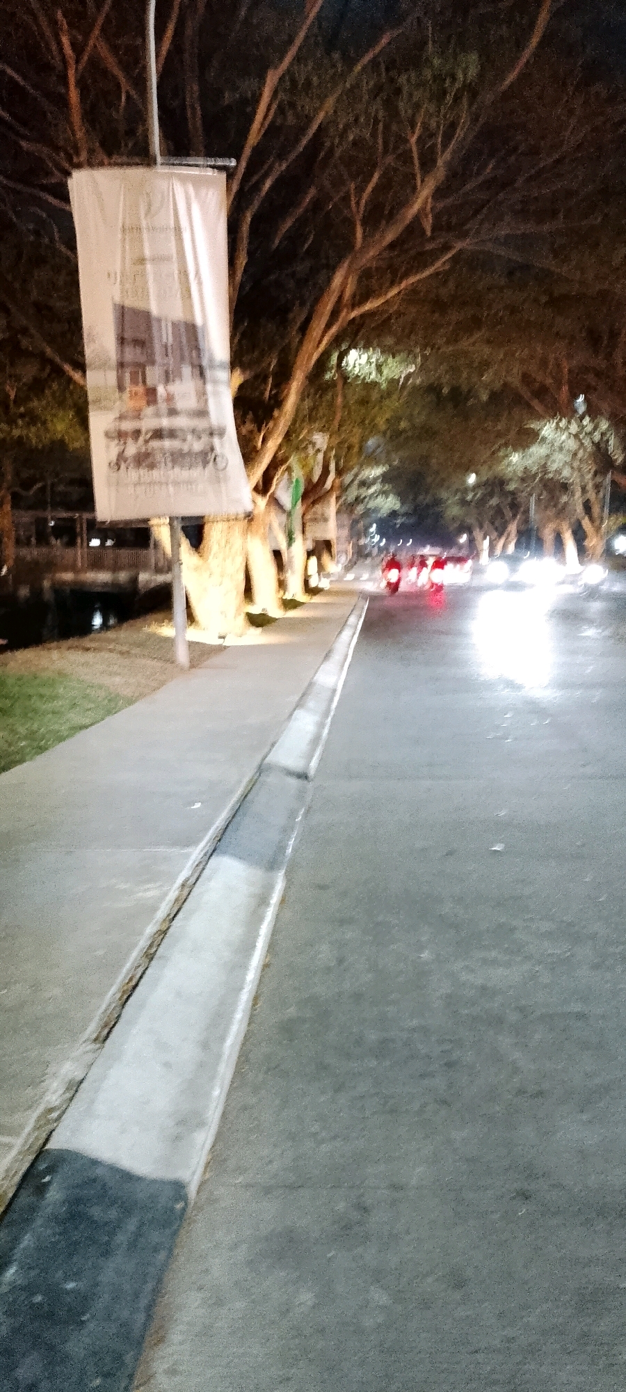 The image depicts a street scene at night in Satriamekar, Indonesia. A paved sidewalk runs along the left side of the road, bordered by a concrete drainage channel. On the sidewalk, a tall, white banner hangs from a pole, featuring an image of a building and text that is partially obscured. Beyond the sidewalk, there are grassy areas and illuminated trees with dense foliage, creating an avenue effect. The road itself is asphalt and appears wet, reflecting the bright headlights of passing vehicles. Several motorcycles are visible on the road, their brake lights glowing red, indicating they are in motion. The overall atmosphere suggests a lively, urban night environment with moderate traffic.