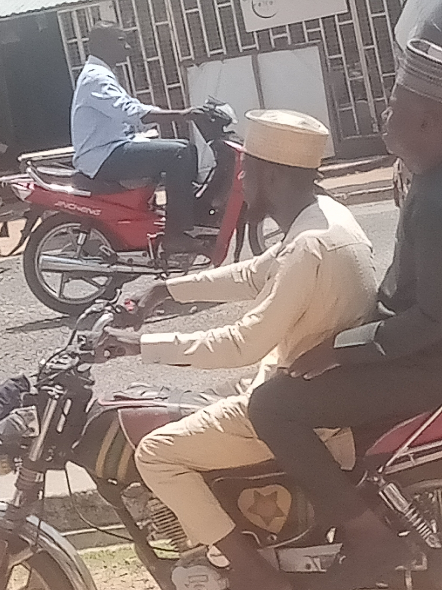 In Jalingo, Nigeria, two individuals are seated on a red motorcycle. The person in the foreground, wearing a beige outfit and a white cap, is positioned in front of the other person. The second individual, partially obscured, is dressed in a dark garment and wears a striped cap. A second red motorcycle, also occupied by a person in a blue shirt, is visible behind them. The scene appears to be taking place outdoors, possibly on a street, with a building featuring barred windows in the background.