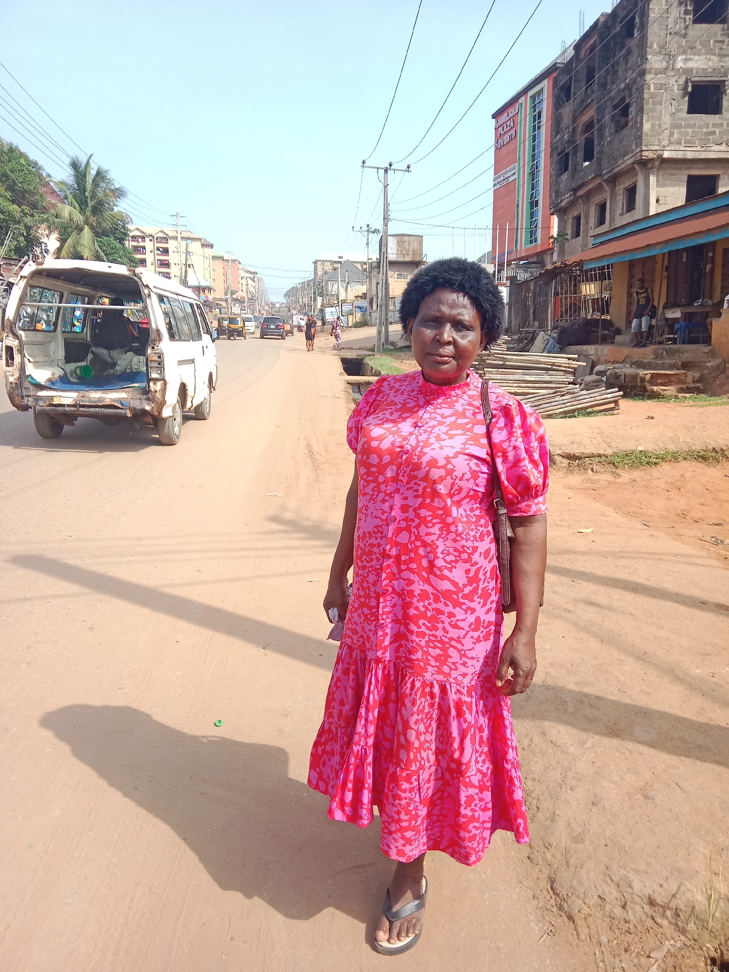 The photo depicts a woman standing on a dirt road in Onitsha, Nigeria. She is wearing a pink and red patterned dress and sandals, and carrying a brown purse over her shoulder. Behind her is a street with vehicles and buildings. A white van with its back door open is visible on the left side of the street. On the right, a partly constructed building and a small shop with people sitting are seen. Power lines stretch across the sky.