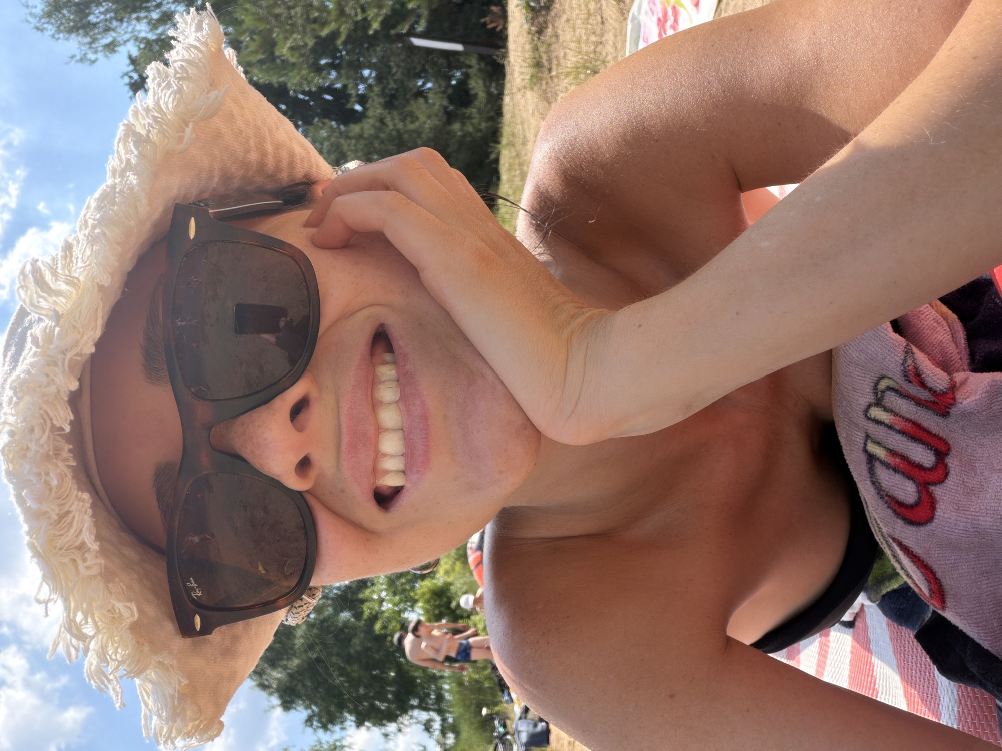 A woman is captured in a close-up, selfie-style photograph, lying down on a beach on a sunny day. She is wearing a wide-brimmed, fringed hat and dark sunglasses. Her head is propped up on her hand, and she is smiling, showing her teeth. Her bare shoulders and upper chest are visible, and she is wearing a black bikini top. A striped towel is beneath her. In the background, there are trees, a body of water, and a few other people, including a man and a child. The overall scene suggests a casual outing at a lakeside or river beach. The image does not contain enough visual information to determine the specific location within Malakhovka, Russia.