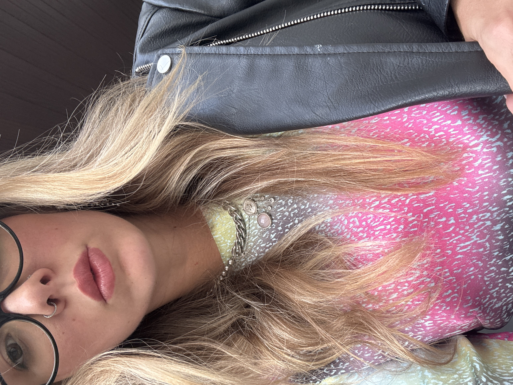 The image shows a close-up, chest-up shot of a woman wearing glasses, a nose ring, a layered necklace with charms, and a black leather jacket over a colorful, patterned shirt. Her blonde hair is partially visible, framing her face. The background is dark and out of focus, suggesting an indoor or dimly lit environment. No specific city or location details are discernible from the image.