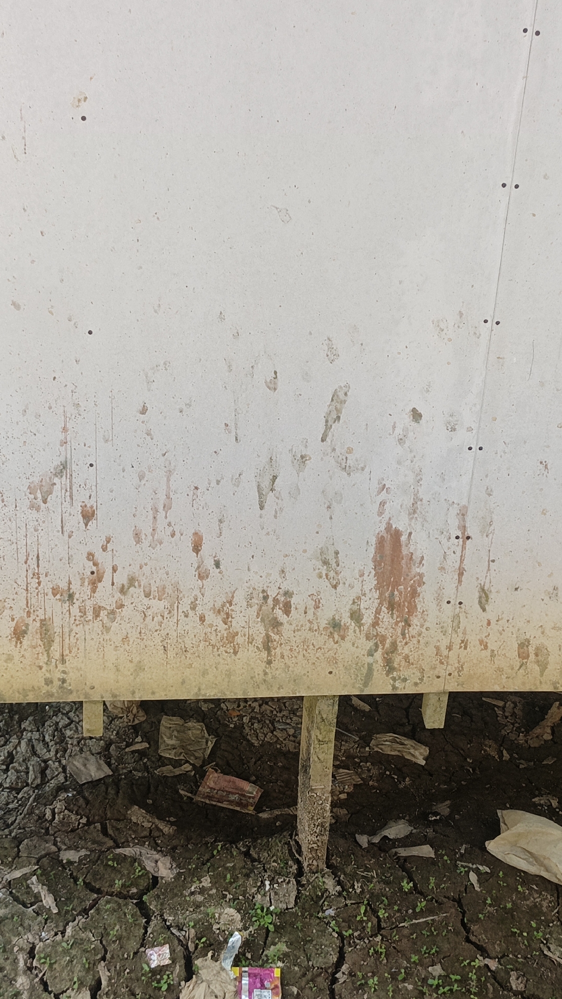 A close-up, vertical shot of a stained, off-white, textured wall with visible dirt and water marks. Below the wall, the scene shows cracked, dry earth with sparse green vegetation. Debris, including pieces of crumpled paper, a red rectangular object, and a white plastic bag, are scattered on the ground. A wooden post is visible, supporting the wall. The image is taken at ground level, looking up at the wall. The location is identified as Sungai Rasau, Indonesia, suggesting an outdoor environment, possibly a rural or underdeveloped area.