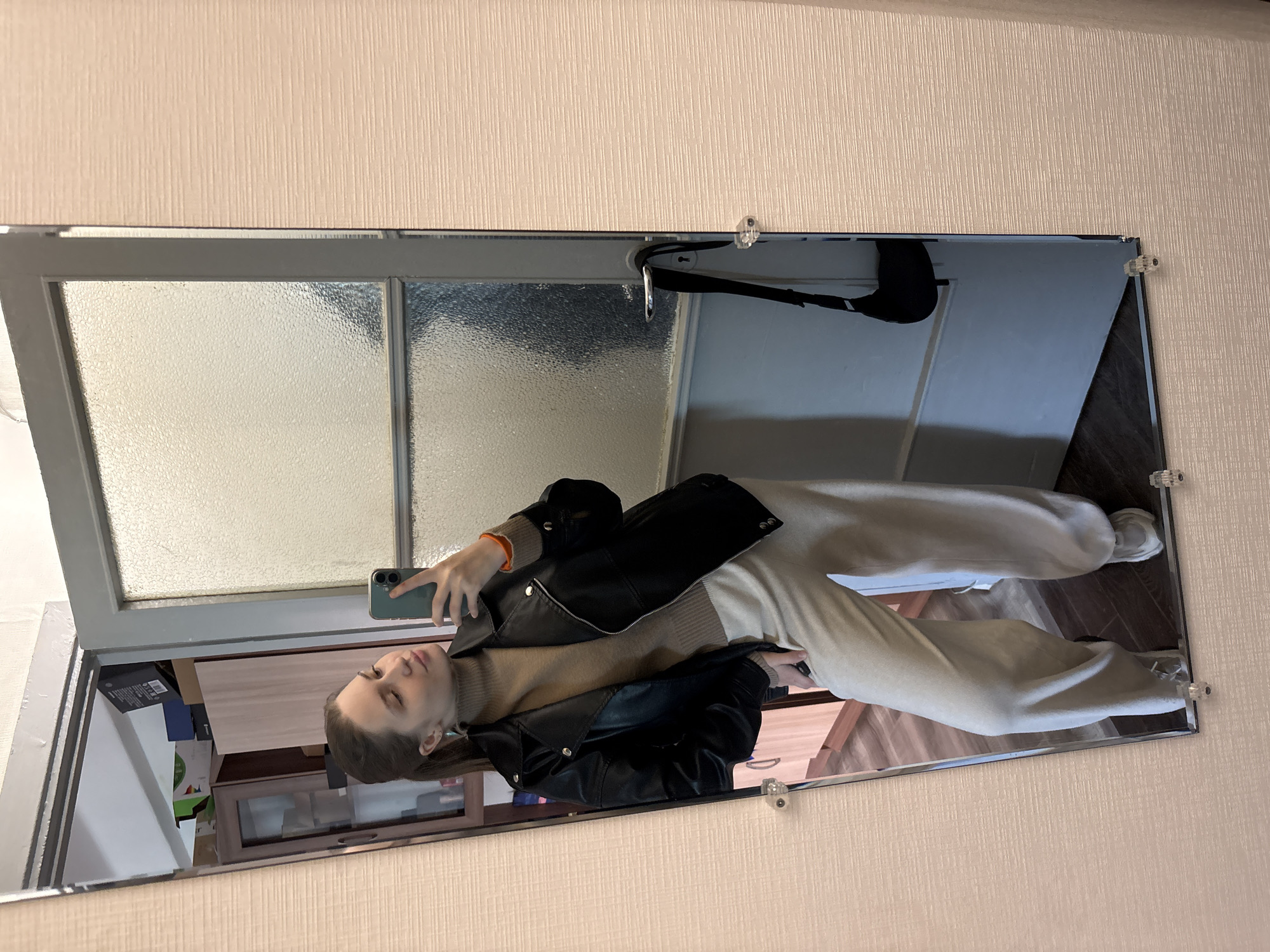 A mirror reflects a woman taking a selfie in Saint Petersburg, Russia. The woman is wearing a black leather jacket, a beige turtleneck sweater, and beige pants. She is holding a green smartphone in her right hand. The reflection shows a portion of a room, including a door with frosted glass, a black handbag hanging on the door, and a cabinet with miscellaneous items. The mirror is attached to a textured wall with clear plastic clips. The floor is visible in the reflection and appears to be dark brown wood.