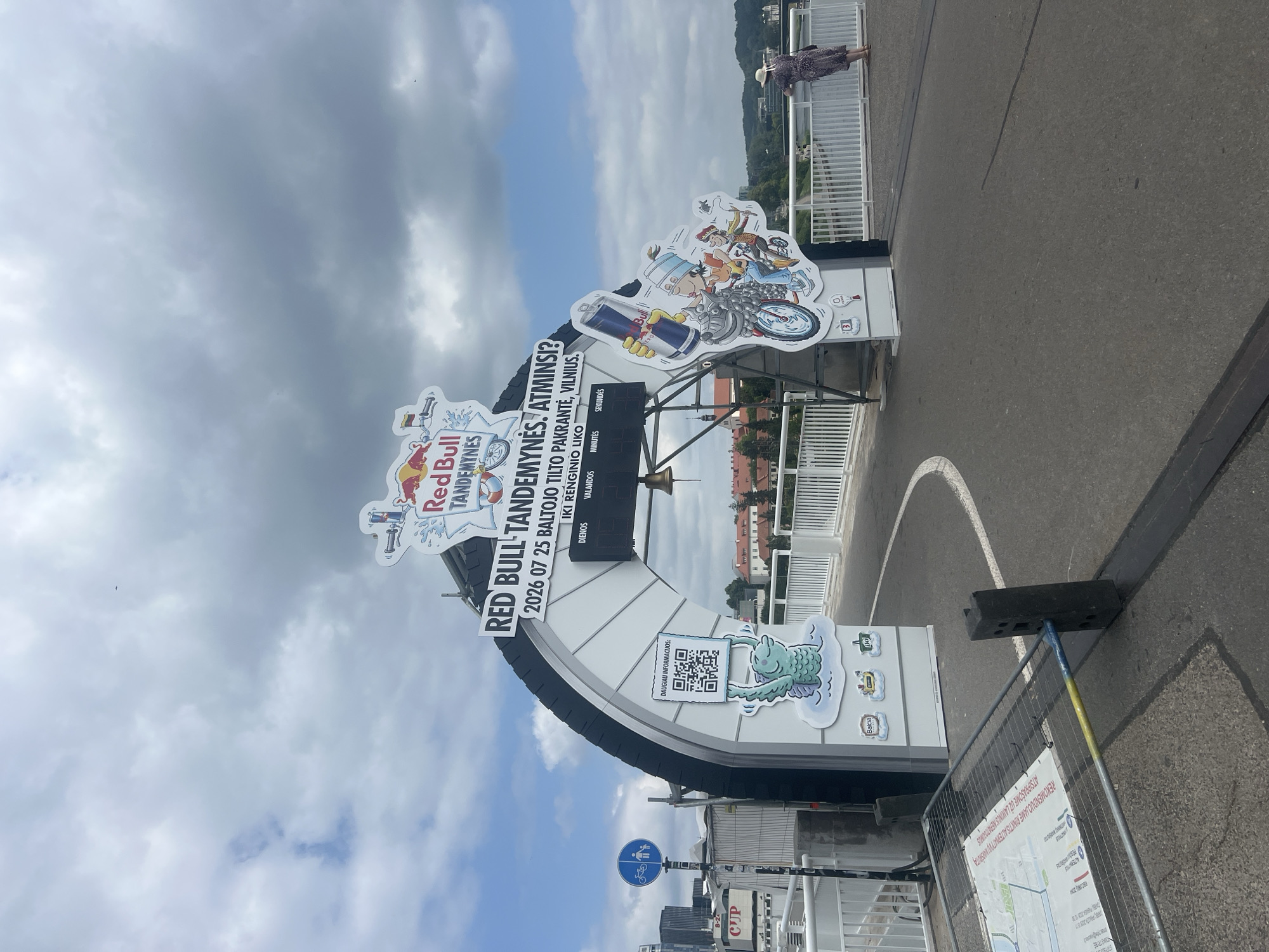 The image shows an arched structure with "RED BULL TANDEMYNES" signage, a countdown timer, and cartoonish graphics on the side. The event appears to be related to Red Bull and possibly tandem activities. A QR code is visible on the left side, along with a drawing of a green creature. On the right side, a graphic depicts people on a motorcycle. The structure is set up on a bridge with a railing, and a body of water and city buildings can be seen in the background. A woman in a patterned dress and a hat is standing on the bridge, looking out at the view. The overall scene suggests an outdoor event or festival in Vilnius, Lithuania. The presence of a countdown timer and event information points to a specific date, "2026 07 25". The setting on a bridge overlooking the city and a river strongly indicates a location within Vilnius.