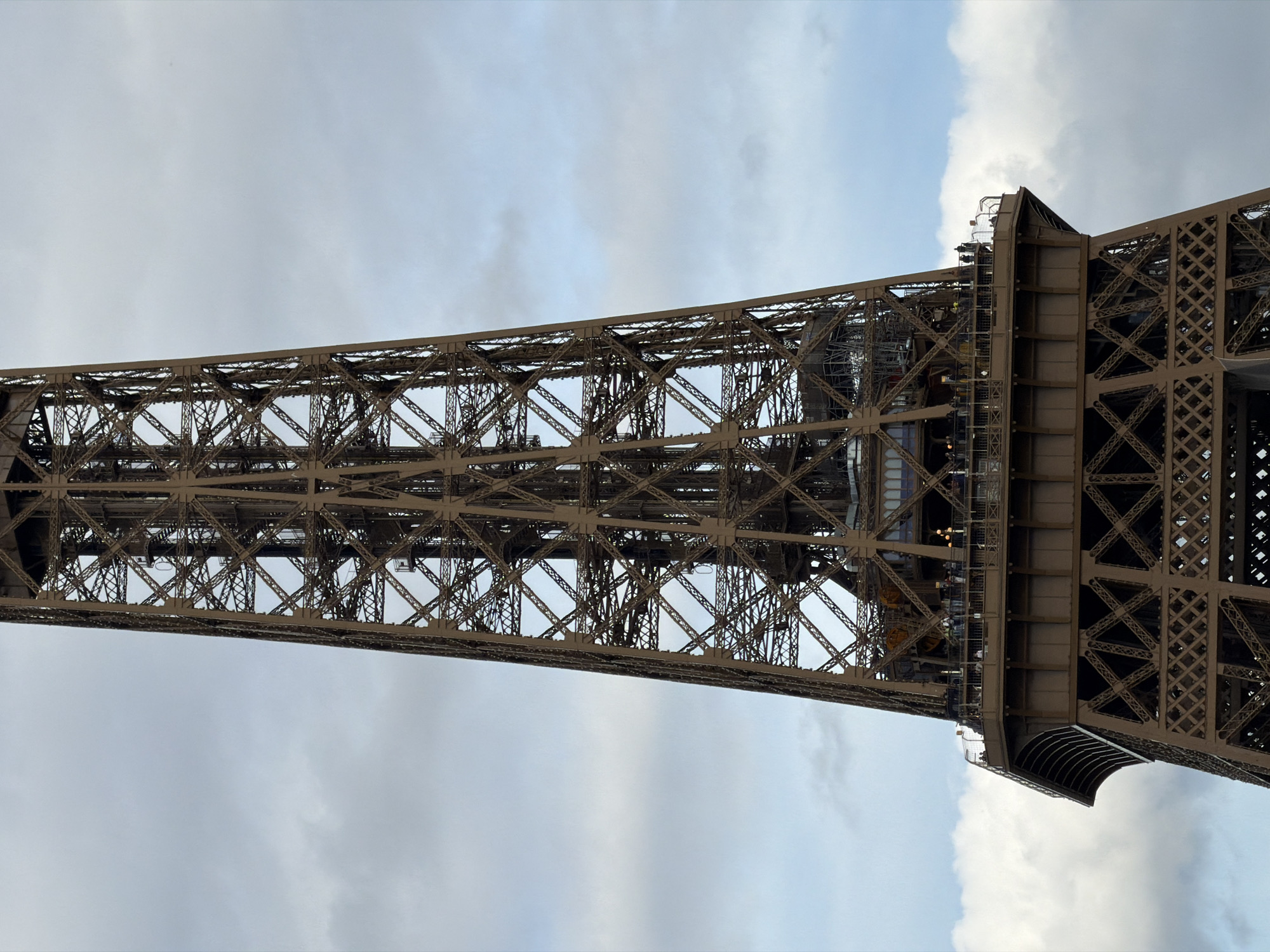 The image shows a low-angle view of the Eiffel Tower in Paris, France. The tower's intricate lattice structure is prominently displayed against a backdrop of cloudy sky. The perspective gives a sense of the tower's immense height and scale. Details of the tower's construction are visible, including beams, rivets, and walkways. The lighting suggests an overcast day. No people are clearly visible in the image, but there is a hint of people on the walkways.