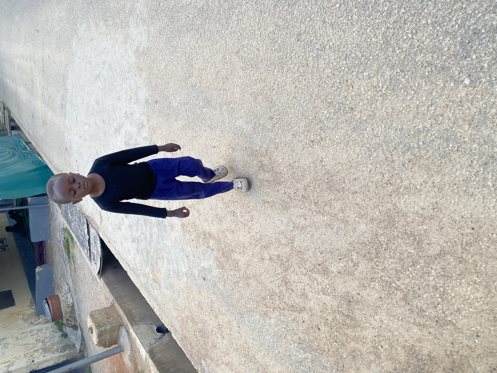 In Jos, Nigeria, a young child walks on a paved pathway adjacent to a drainage channel. The child, dressed in a black long-sleeved shirt and blue pants, wears white clogs with a cartoon character. The pathway is made of light-colored concrete aggregate. To the left of the pathway is a concrete drainage channel with some debris. Behind the channel, a weathered building with a yellowish facade is visible, with another structure partially obscured. In the background, a green tarp is stretched over a frame, and a person is seated on the ground. The scene suggests an outdoor urban or semi-urban environment.