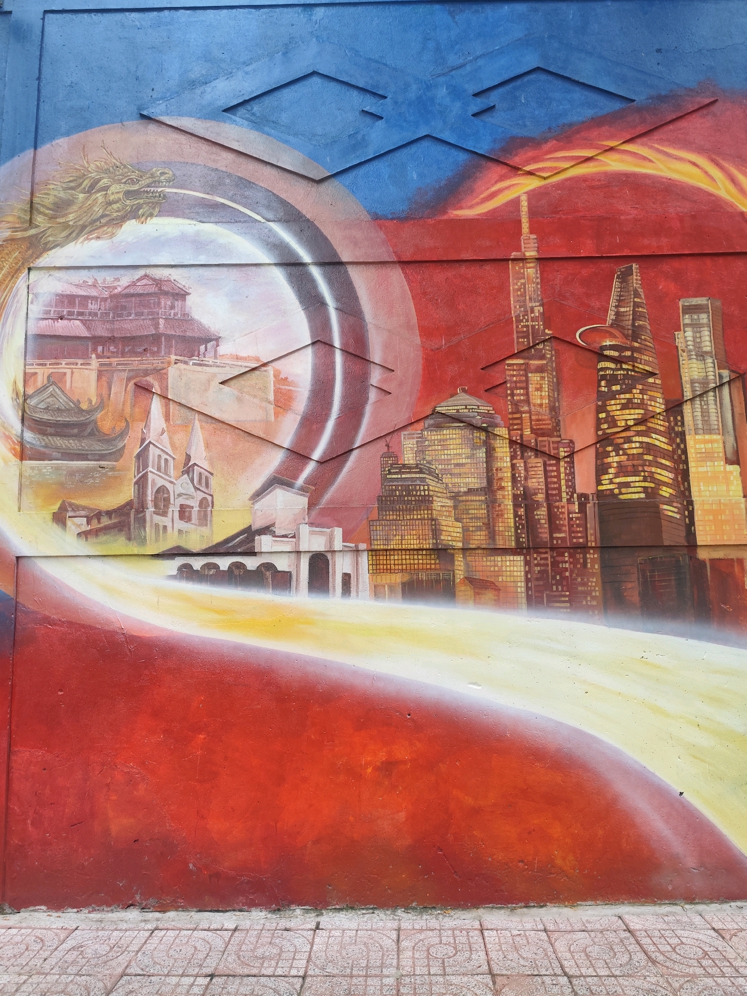 The image is a photograph of a mural painted on a concrete wall in Ho Chi Minh City, Vietnam. The mural depicts a blend of traditional and modern elements. On the left side, a golden dragon is painted, breathing a stream of water or mist. Behind the dragon are traditional Vietnamese buildings with tiered roofs. In the center and to the right, a modern cityscape is portrayed with tall skyscrapers lit up at night. The mural is divided into sections by geometric shapes and diagonal lines, with a large red and yellow swirling design at the bottom and a blue section at the top with abstract diamond shapes. The wall appears to be located outdoors, as evidenced by the tiled sidewalk in the foreground. The overall impression is a vibrant and symbolic representation of the city's heritage and progress.