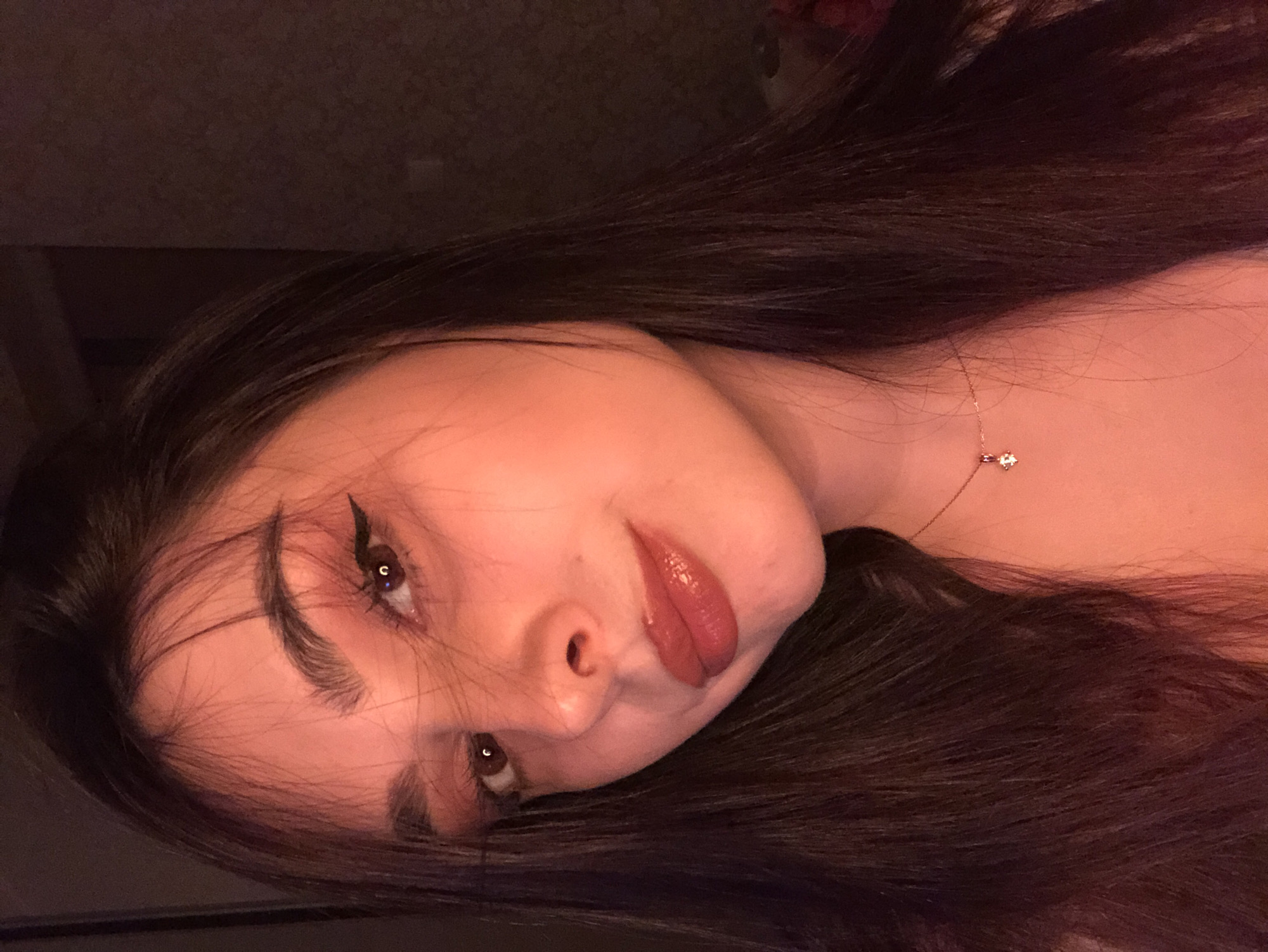 A young woman with long, dark hair is pictured indoors with a tilted perspective. She has fair skin, dark eyes, winged eyeliner, and glossy lips. She is wearing a delicate necklace with a small pendant. The background is dark and indistinct. No information suggests the city of Noviy, Russia.