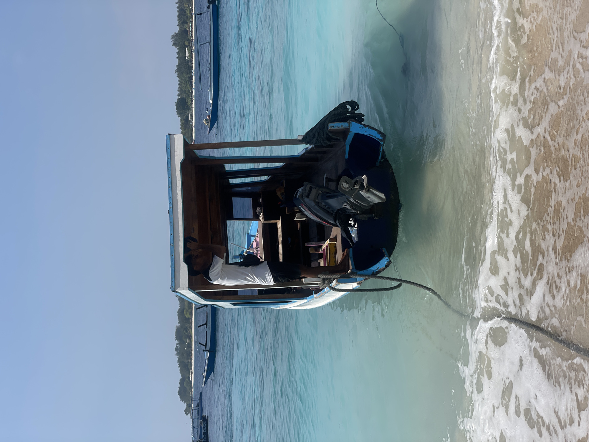 An eye-level medium shot captures a small blue and white boat partially submerged in clear turquoise water near a sandy beach. The boat is tethered to the shore by a thick rope that is visible partially submerged in the water and trailing onto the sand where gentle waves are creating foam. A person in a white t-shirt and black shorts is standing on the boat's deck, holding a black object to their face as if looking through binoculars. The boat has a wooden cabin structure with an outboard motor attached to the stern. In the background, several other boats are anchored in the calm water, and a lush green island with trees is visible on the horizon under a clear blue sky.