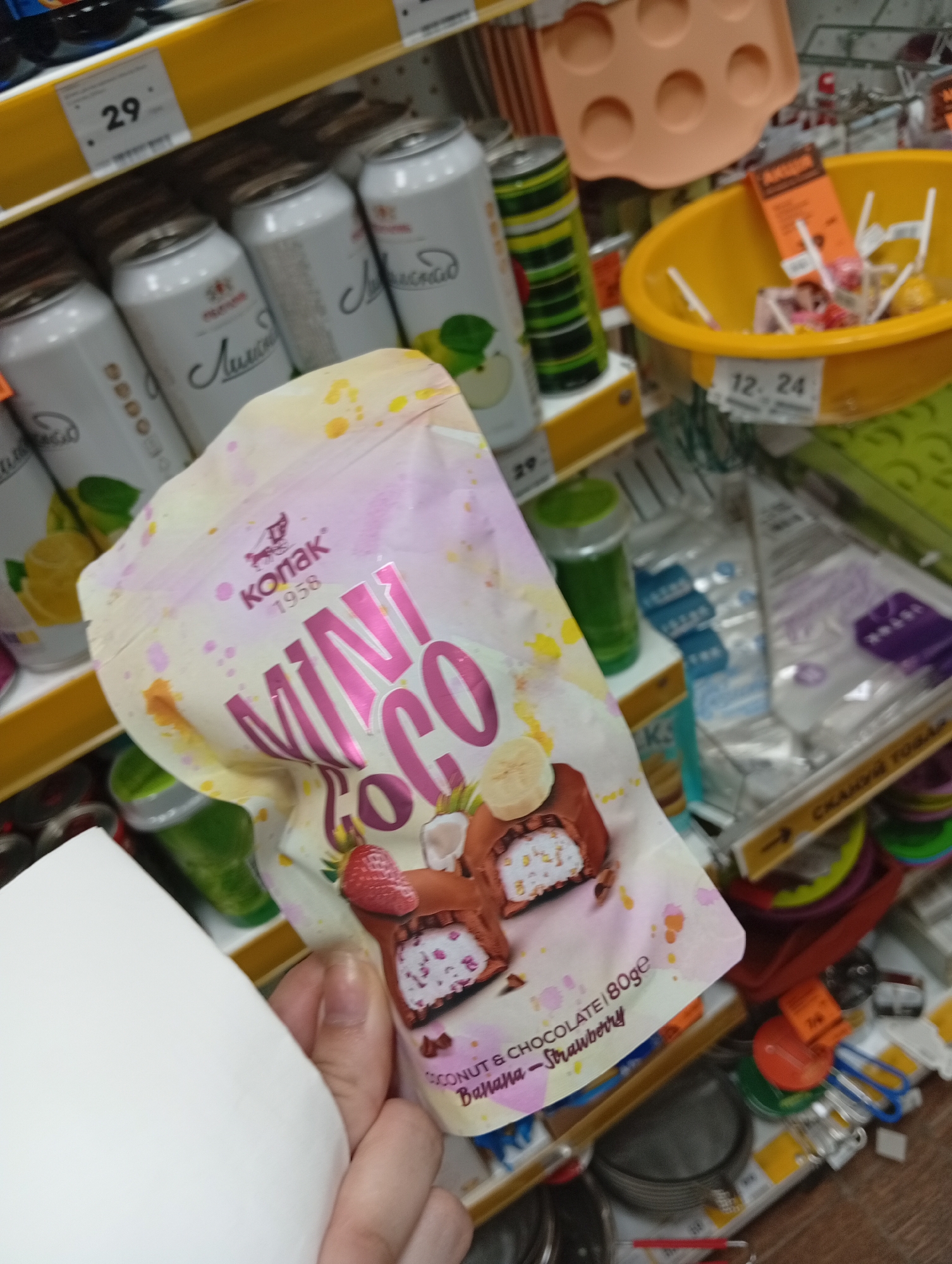 The image shows a close-up of a hand holding a package of "MINI COCO" candy. The candy package is pink and white with colorful splatter designs. The text on the package indicates it is made of coconut and chocolate, with banana and strawberry flavors. The background is blurred and shows a shelf in what appears to be a store. On the shelf are various items including cans of drinks, a yellow bowl with candy, and other packaged goods. The overall scene suggests a retail environment, likely a supermarket or convenience store. The provided context specifies the location as Poltava, Ukraine.