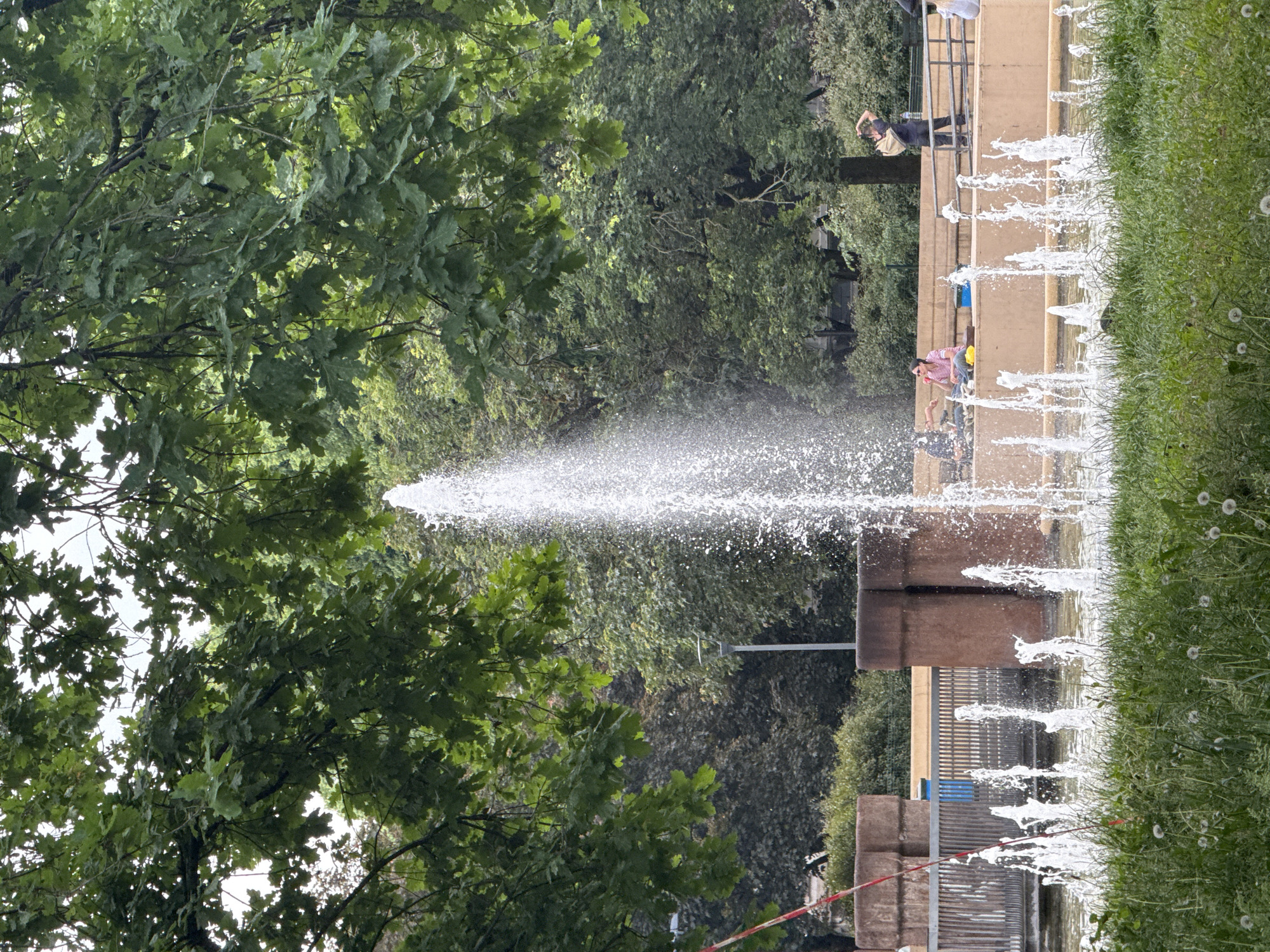 The image captures a vibrant outdoor scene in what appears to be a public park or square in Milan, Italy. A prominent fountain dominates the center and right side of the frame, featuring a powerful central jet of water spraying high into the air, surrounded by numerous smaller, rhythmic jets lining its basin.
To the left, a dense canopy of lush green tree foliage fills a significant portion of the image. On the right, a verdant strip of grass, dotted with small white dandelion-like flowers, runs alongside the fountain's edge.
In the mid-ground, behind the fountain, several people are visible. One individual wearing a light pink or striped shirt and dark pants stands near the fountain, seemingly observing it. Further back, another person in a dark top and light pants is visible near a metal railing. Other figures are present but less distinct. The fountain structure itself is composed of brown, block-like stone or concrete elements. A red and white striped barrier tape is partially visible in the lower right portion of the frame, stretching across the base of a fountain structure.
The scene takes place during the daytime, characterized by soft, diffused lighting, suggesting an overcast sky or an area with dappled light. The overall mood appears peaceful and leisurely, with people enjoying the public amenity. No discernible text is visible in the image.