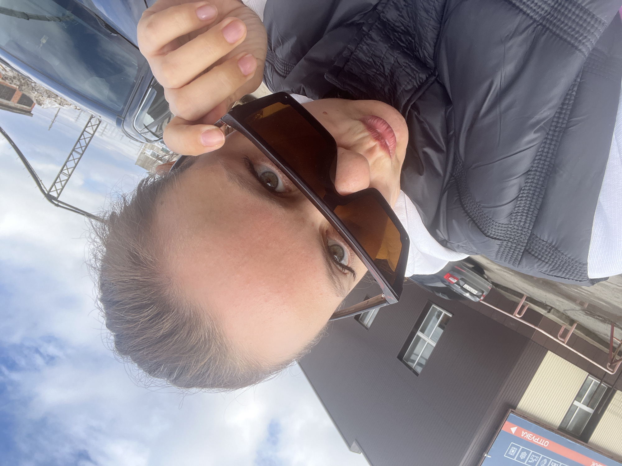A person is holding sunglasses, looking into the camera. They're outdoors, in what appears to be the city of Chaikovsky, Russia. The scene includes a building with windows, a car parked in the background, and a sign in Russian, indicating this is likely a public facility. The person wears a black jacket. The sky is visible with some cloud cover.