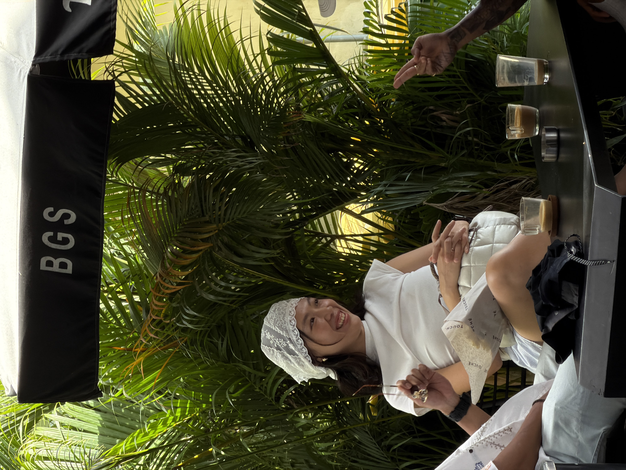Here's a description of the image:
The photograph shows a woman with dark hair wearing a white headscarf and a white shirt, smiling and looking toward the upper right. She is holding what appears to be a small object in her right hand. Next to her, on a black surface, are three glasses with liquid in them, possibly for a beverage tasting. A man's hand with a tattoo is pointing towards the glasses. Behind the woman and the table is dense foliage, likely tropical plants. A black banner with white text that reads "BGS" is visible on the left side of the image. Based on this limited visual evidence, it's difficult to ascertain whether this image was taken in the city of North Kuta, Indonesia. More information is needed.