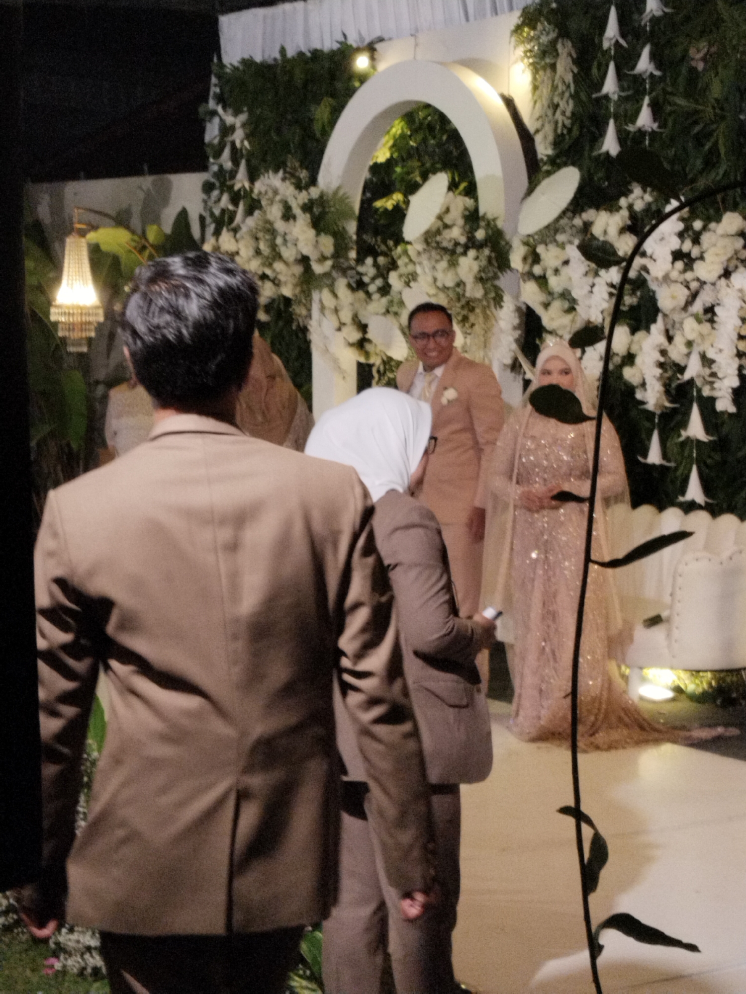 The image captures a wedding ceremony, likely in Serang, Indonesia, given the context. In the foreground, a person in a brown suit is facing away from the camera, their back to the viewer. To their right, another individual, also in a brown suit, is partially visible. The central focus of the image is a bridal couple. The groom, wearing a peach-colored suit and glasses, stands to the left of the bride. The bride is adorned in a sparkling rose-gold gown and a matching veil, her head covered. They are positioned in front of an elaborate floral backdrop featuring white flowers and lush greenery, arranged around a white archway. To the far left, a decorative lamp with multiple tiers of lights hangs, illuminating the scene. The overall setting suggests an indoor or covered outdoor venue decorated for a formal event.
