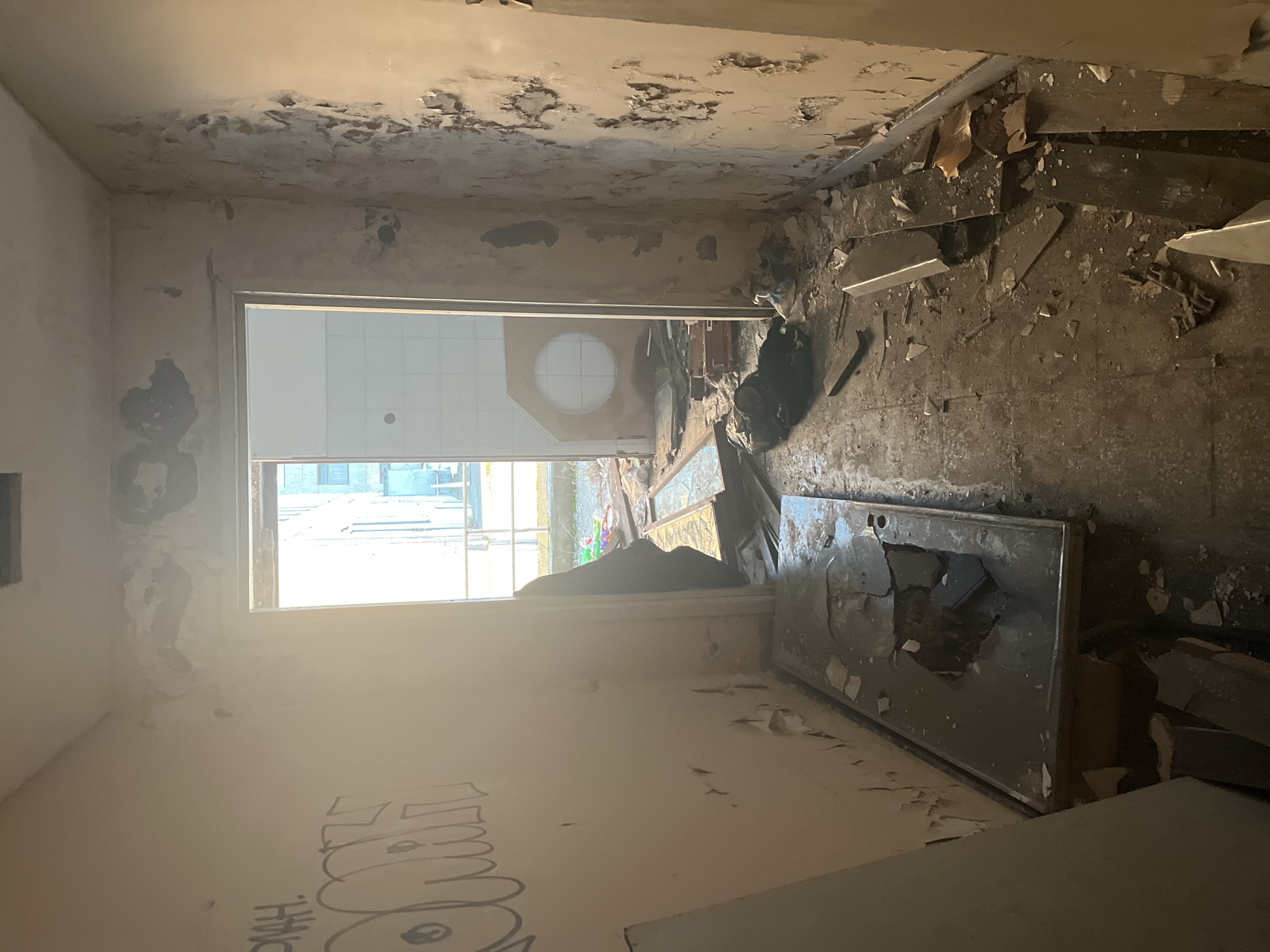 The image depicts the interior of a severely dilapidated building. The location, Марковский, Russia, is not confirmed through visual elements. The scene shows significant deterioration: peeling paint, damaged walls and ceilings, and debris strewn across the floor. A broken door or panel lies on the floor, with a large hole. Graffiti is visible on the wall in the foreground. Through a doorway, there's a view of another room with a window revealing an exterior scene with buildings. No people or subjects are visible. The environment is characterized by neglect and decay.