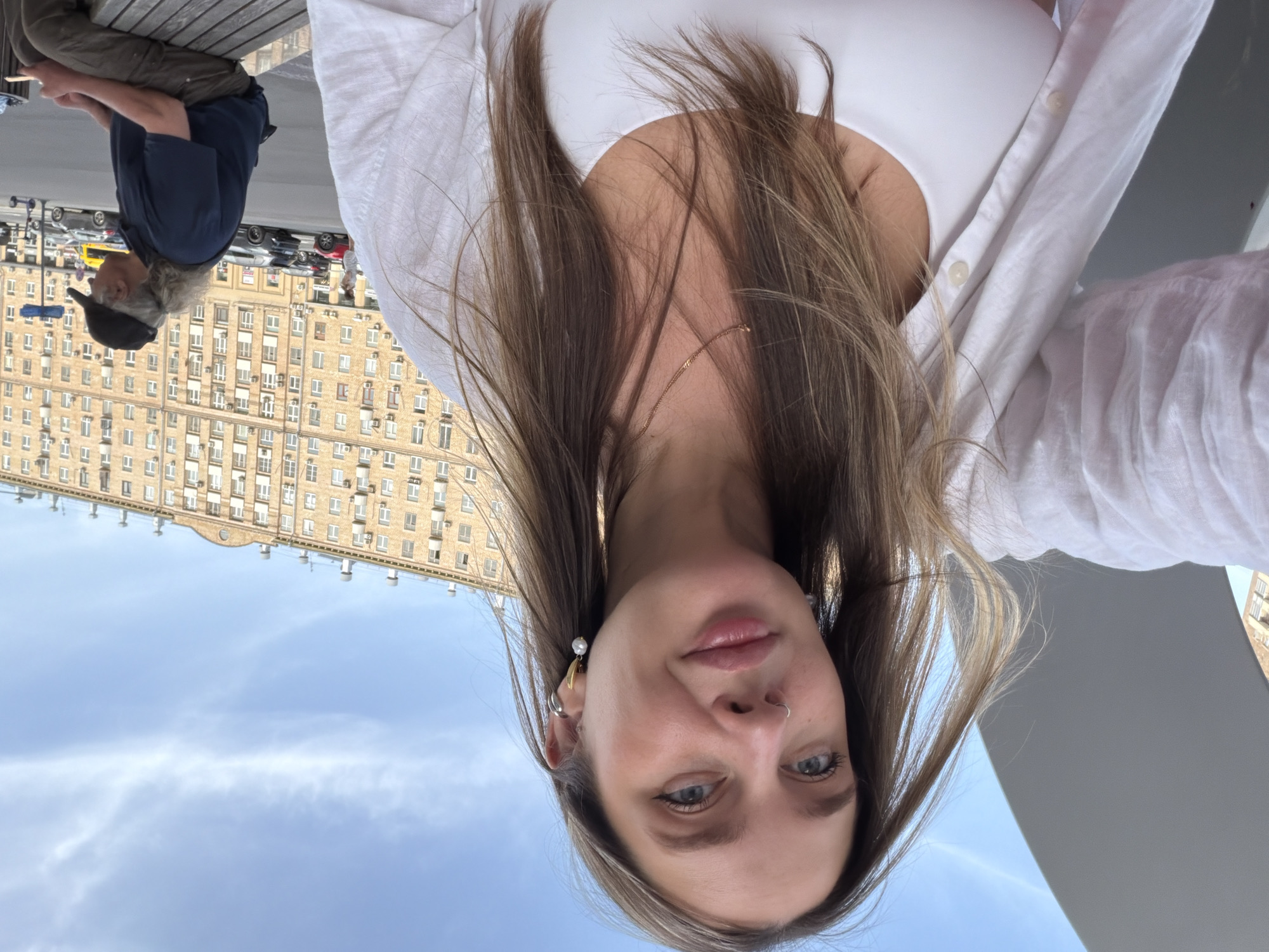 A rotated eye-level shot shows a fair-skinned person with long brown hair wearing a white top. They are outside in Moscow, Russia. In the background, there is a large, multi-story, light-colored building with many windows. A man wearing a navy blue shirt and a dark cap is seated to the left. Cars line the street. A light blue sky is visible.